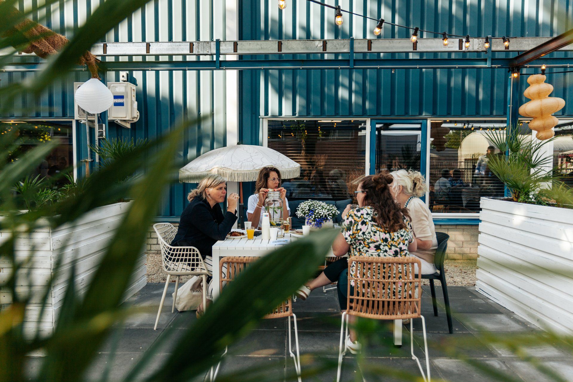 Een groep mensen zit buiten aan een tafel te praten en te eten, in de omgeving van een blauw gebouw.