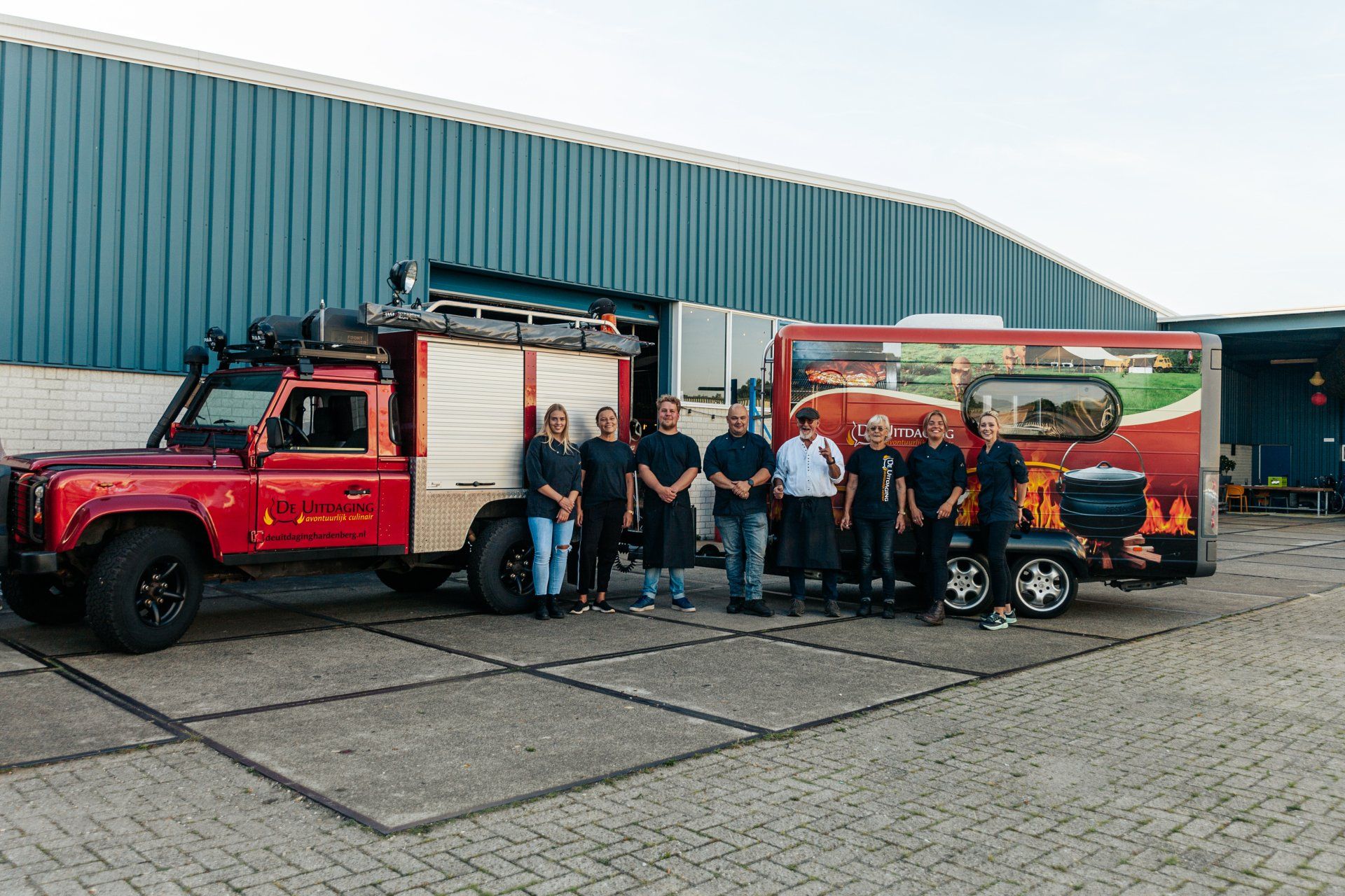 Rode vrachtwagen met aanhanger; een groep mensen poseert ervoor. Gebouw op de achtergrond.