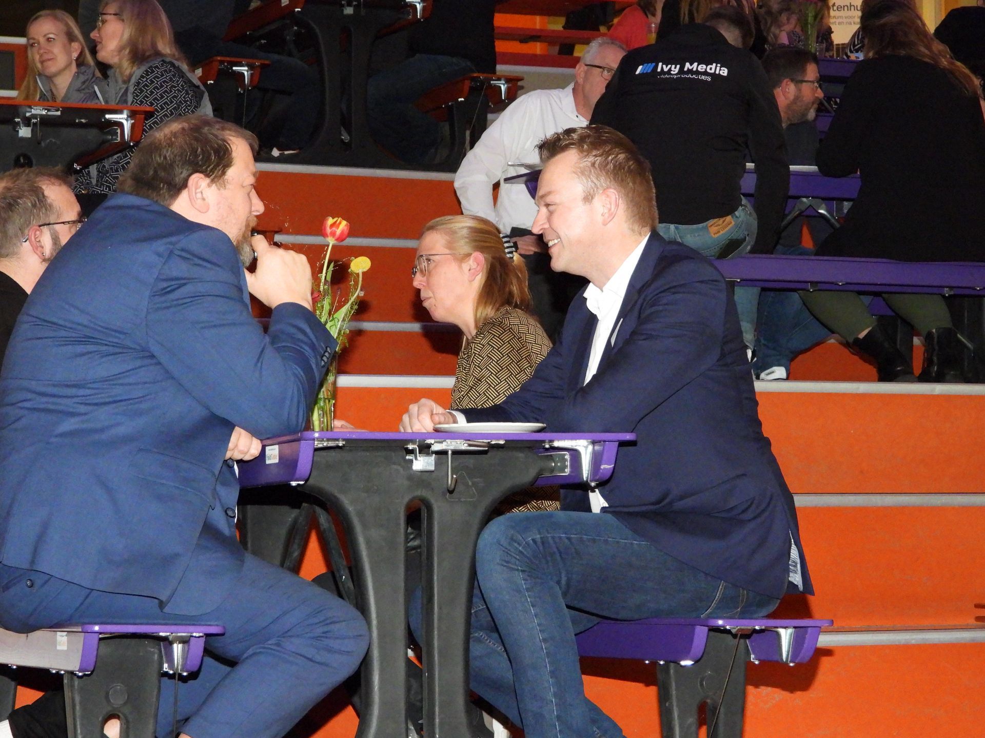 Twee mannen in pak zitten aan een tafel en voeren een gesprek in een drukke, felverlichte ruimte.