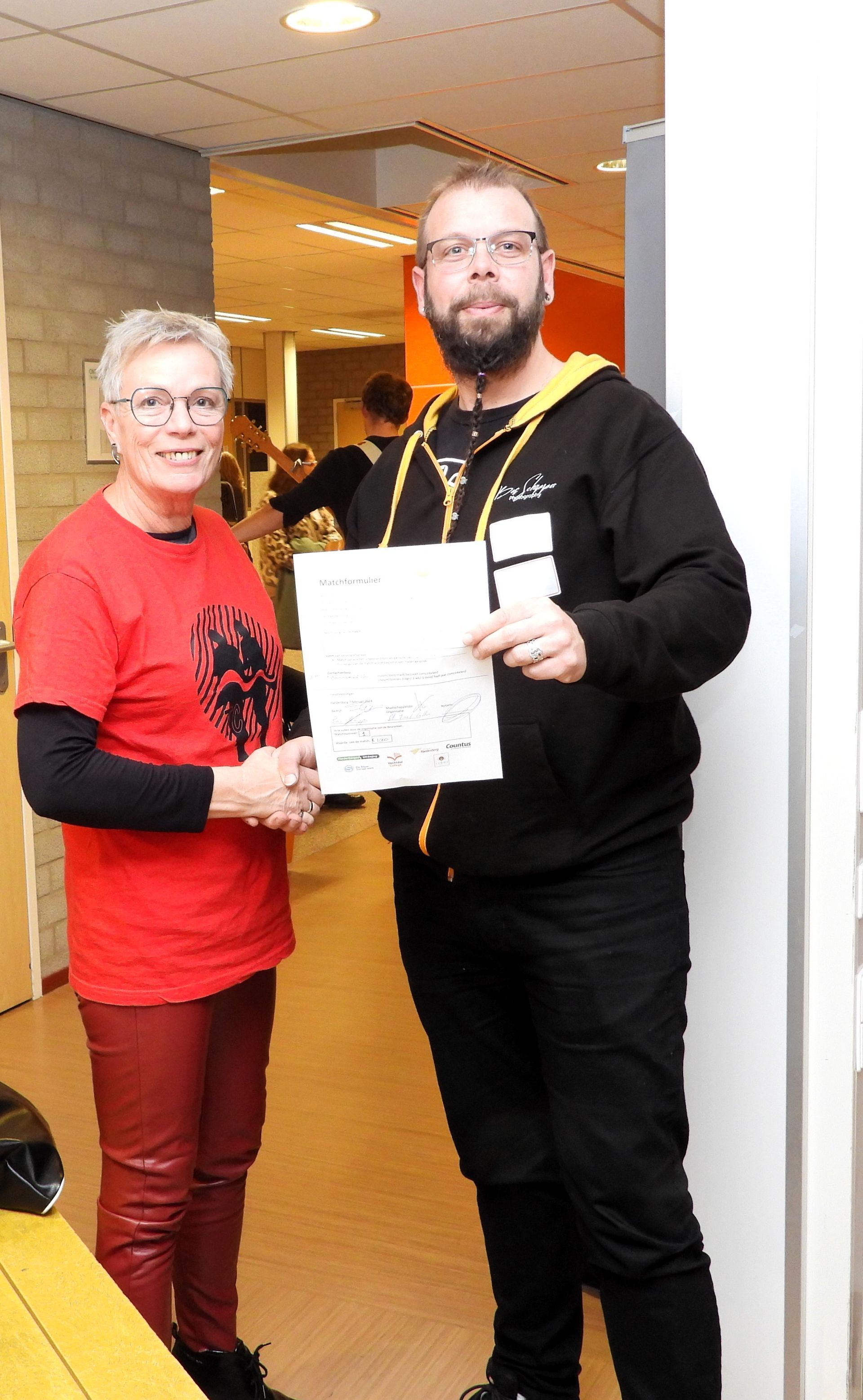 Vrouw in rode blouse en leren broek met man in zwart die papier vasthoudt; binnen.