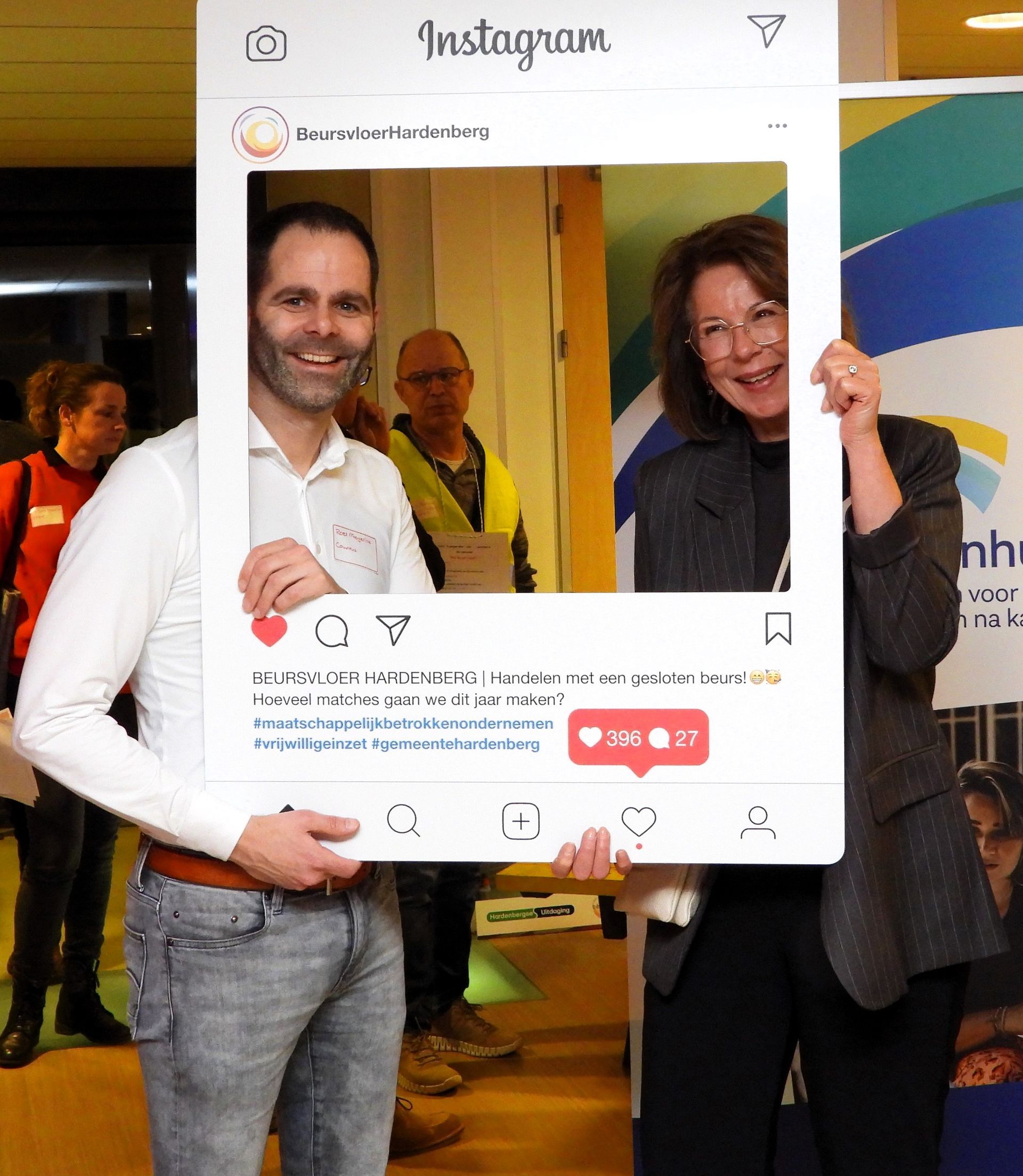Twee mensen poseren binnen met een Instagram-frame. De man lacht in een wit overhemd en spijkerbroek. De vrouw lacht met een bril en een blazer aan.
