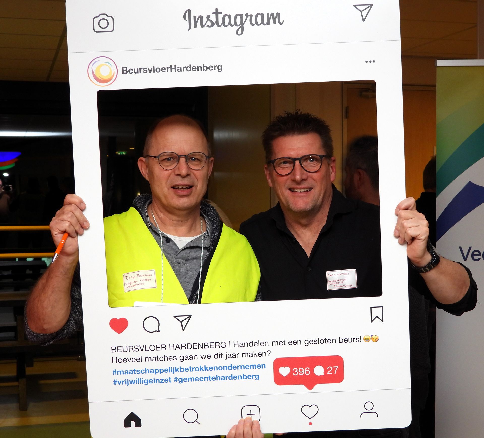 Twee lachende mannen poseren voor een Instagramfoto. De ene draagt ​​een geel vest, de andere een zwart shirt.