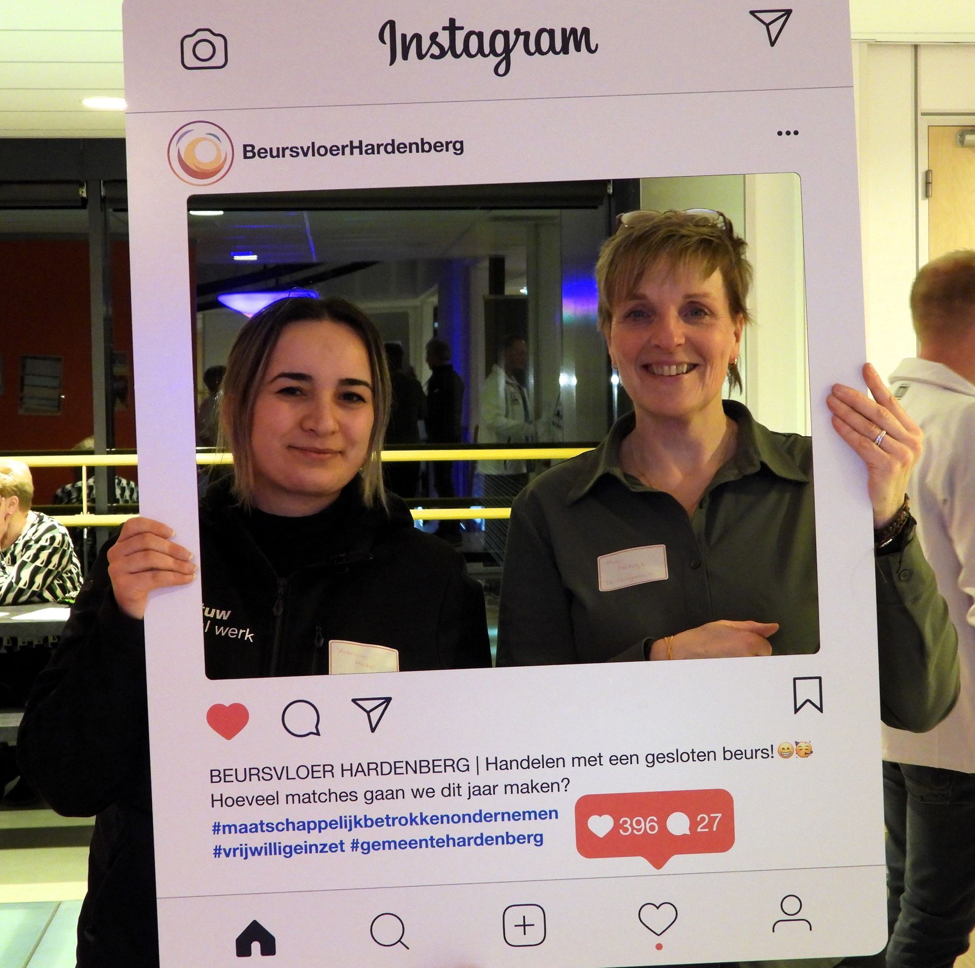 Twee vrouwen poseren lachend met een Instagram-fotolijst. Een van hen houdt de lijst vast, die een moderne kantooromgeving afbeeldt.
