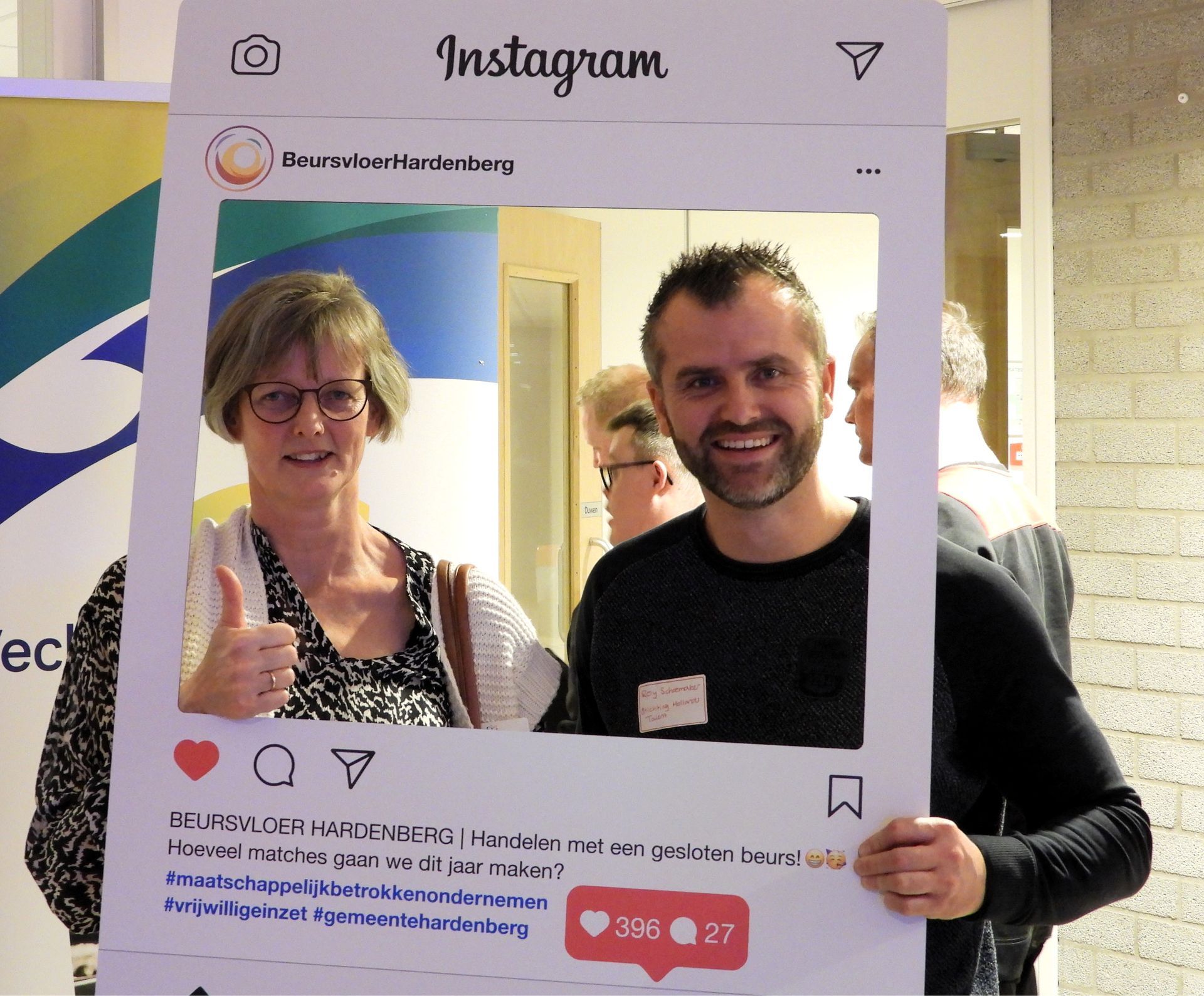 Twee lachende mensen poseren voor een Instagramfoto tijdens een evenement; de vrouw steekt haar duim omhoog.