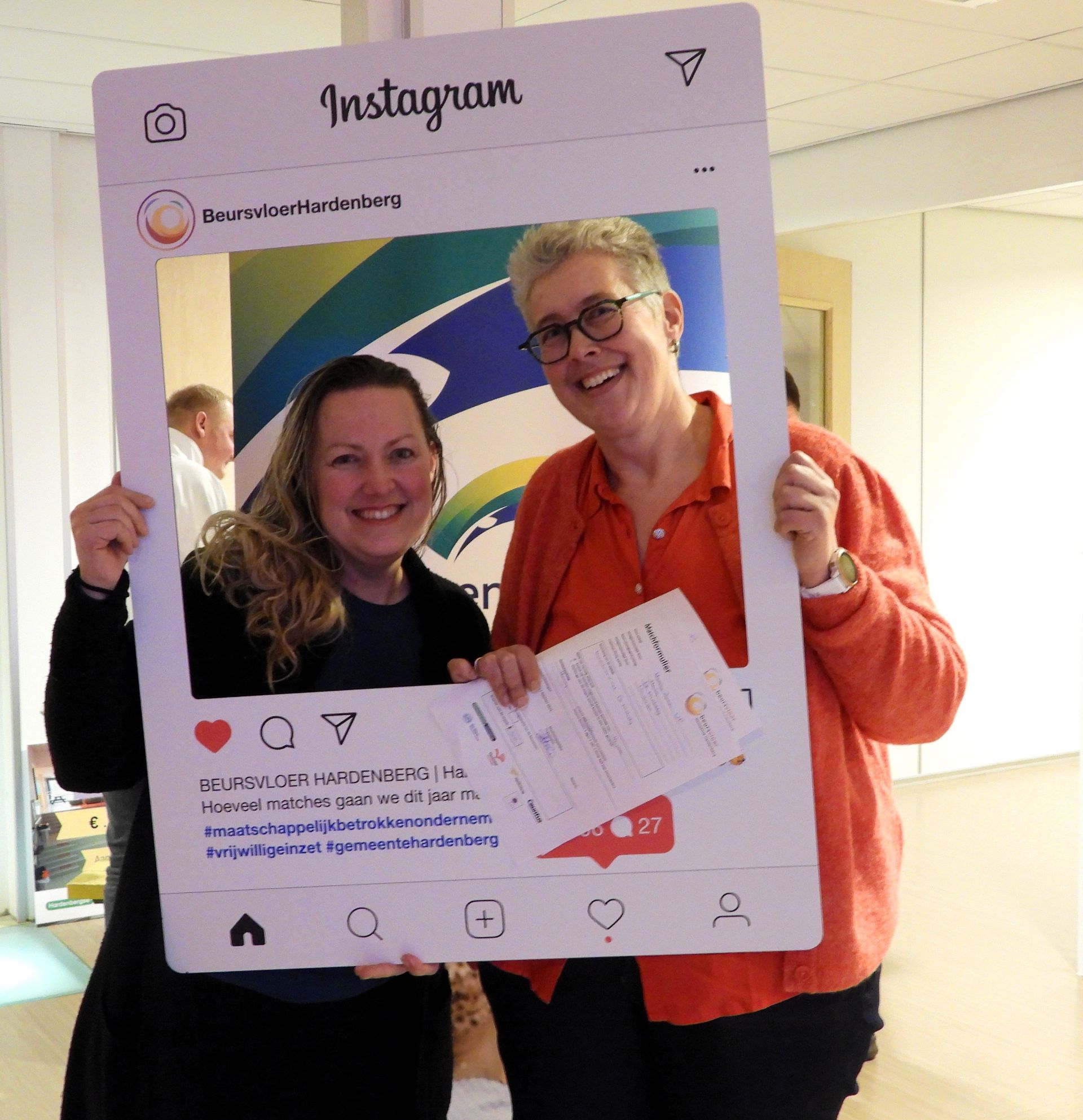 Twee vrouwen glimlachen en houden een Instagram-fotolijstje vast. Ze staan ​​voor een spandoek.