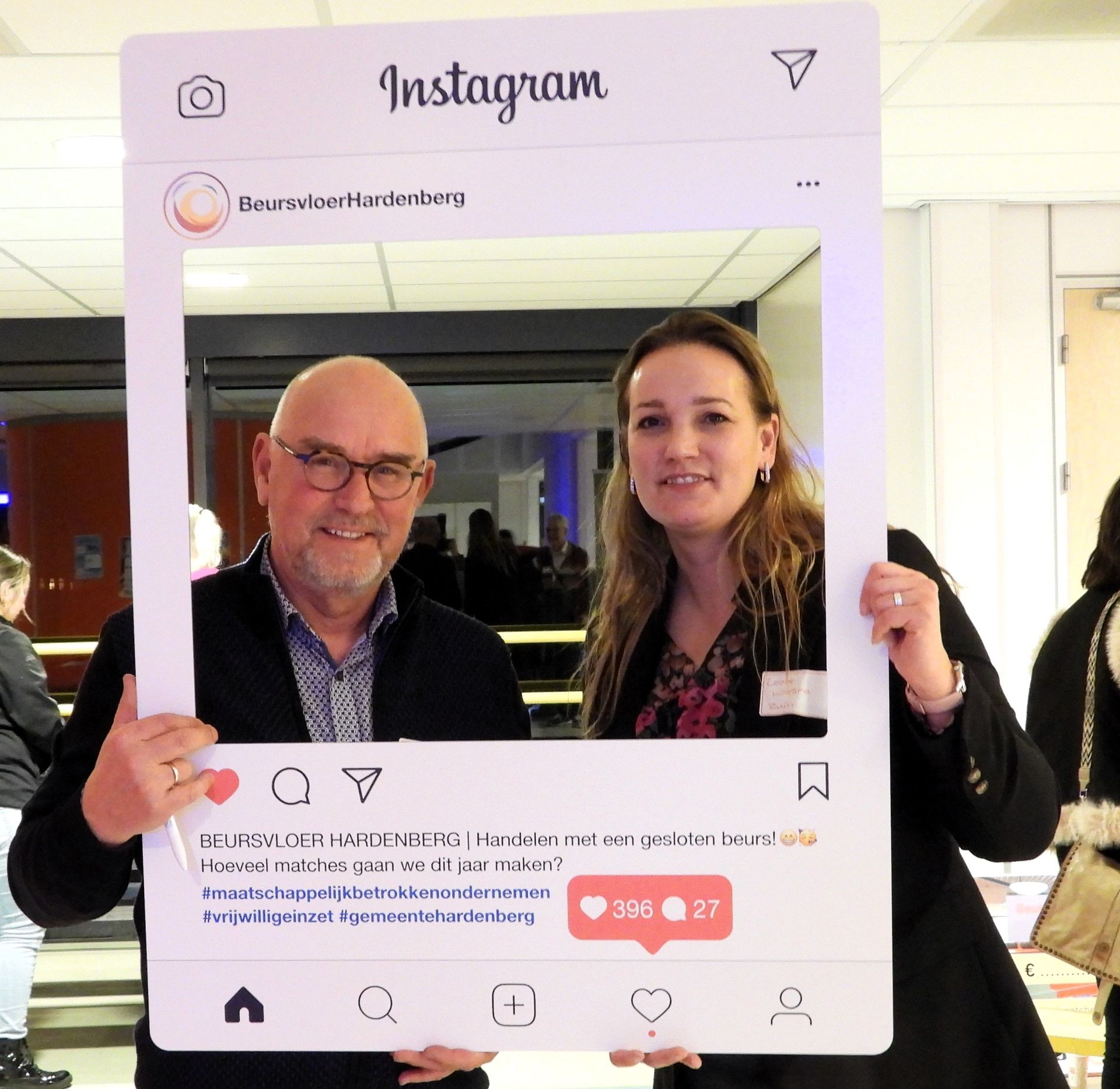 Twee mensen poseren met een oversized Instagram-fotolijst. De man draagt ​​een bril en een blazer. De vrouw lacht. Binnen.