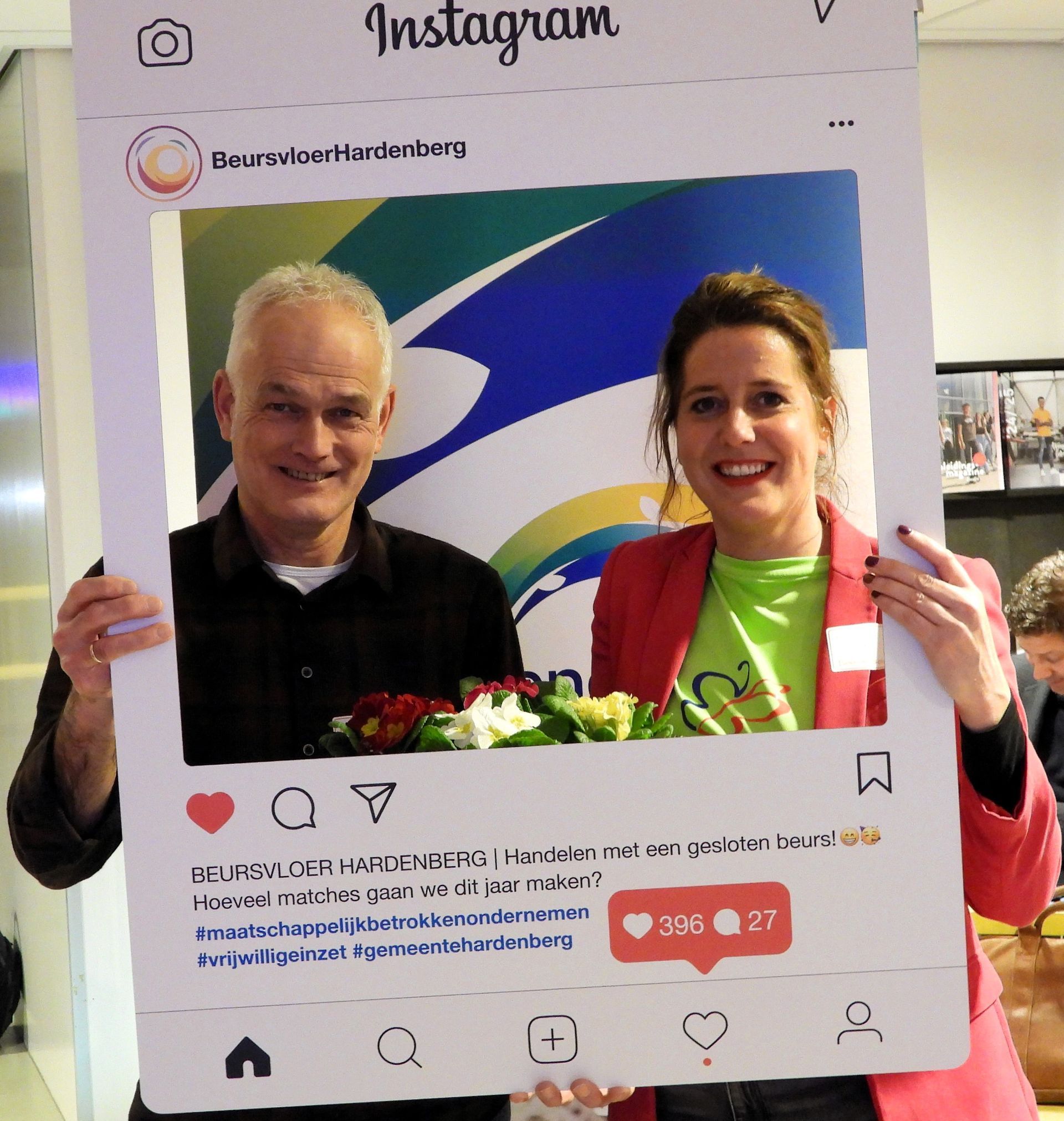 Twee mensen poseren in een Instagram-frame. Een man in een zwart shirt, een vrouw in een rode blazer. Op de achtergrond is een kleurrijk logo te zien.
