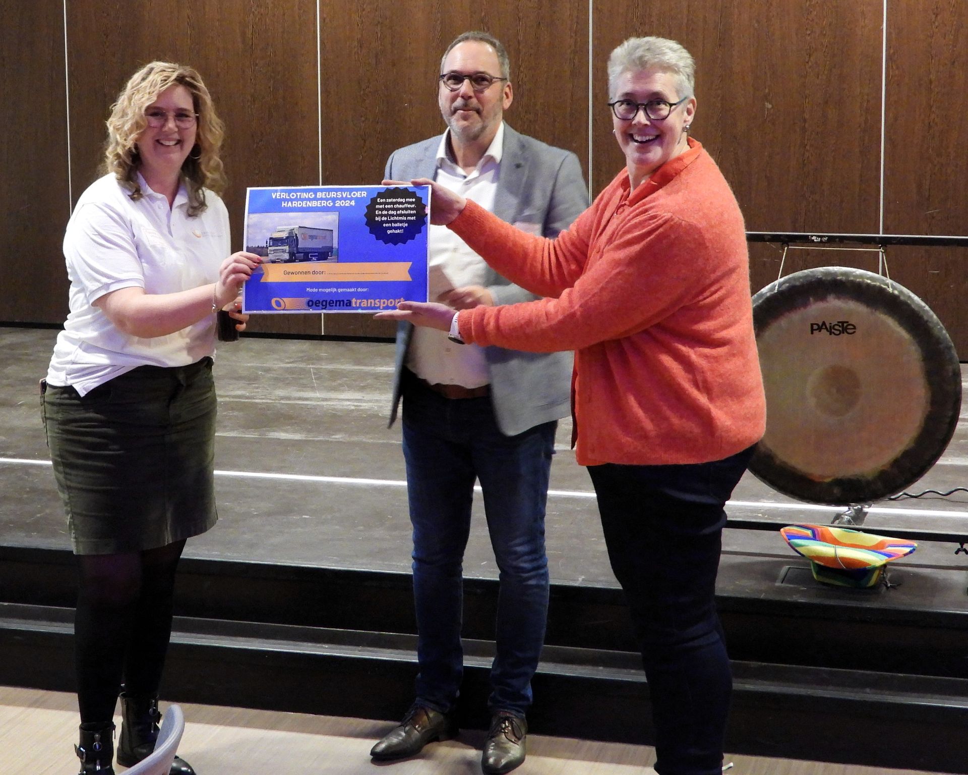 Drie mensen staan ​​met een cheque in hun handen in een auditorium en glimlachen. Een van hen draagt ​​een oranje shirt. Een gong staat er vlakbij.