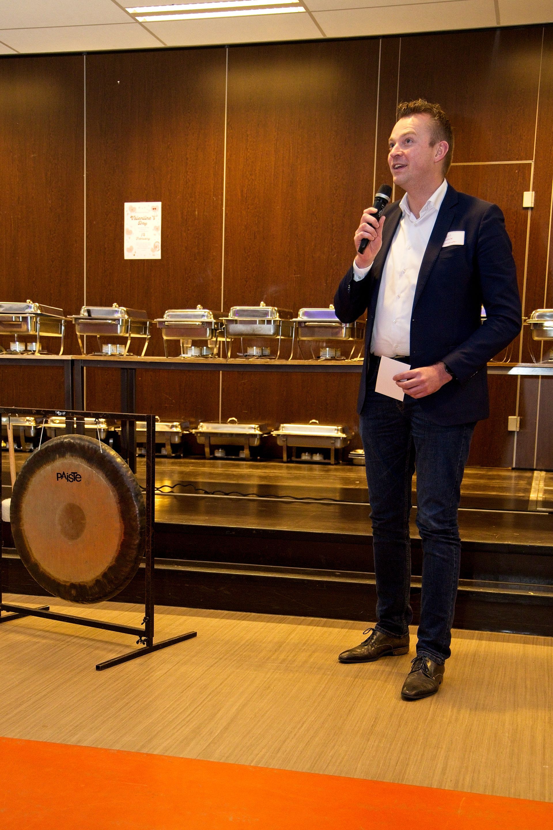 Een man in pak spreekt in een microfoon tijdens een evenement met een buffet en een gong.
