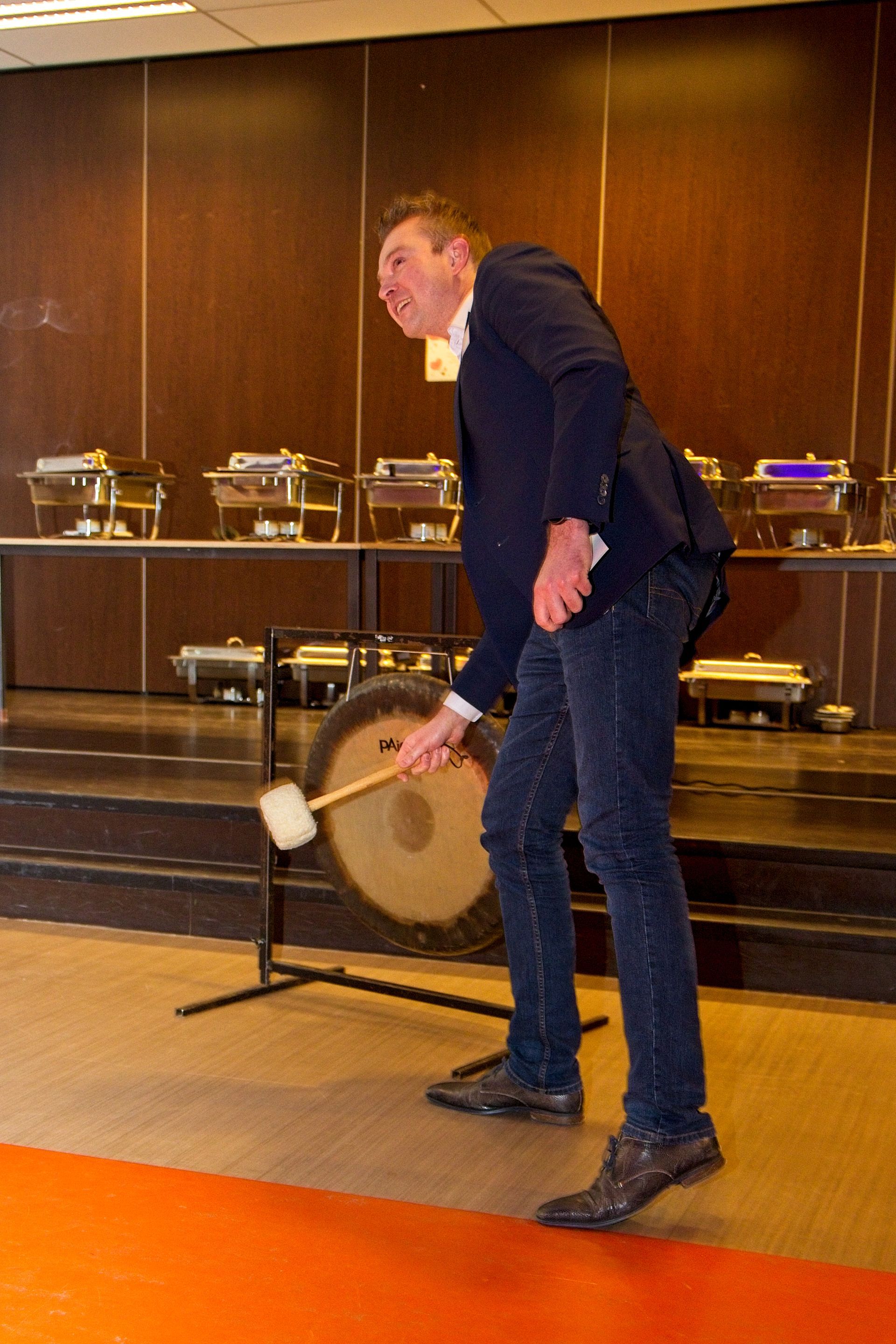 Een man slaat met een hamer op een grote gong. Hij draagt ​​een blazer en een spijkerbroek en staat vlakbij een buffet.