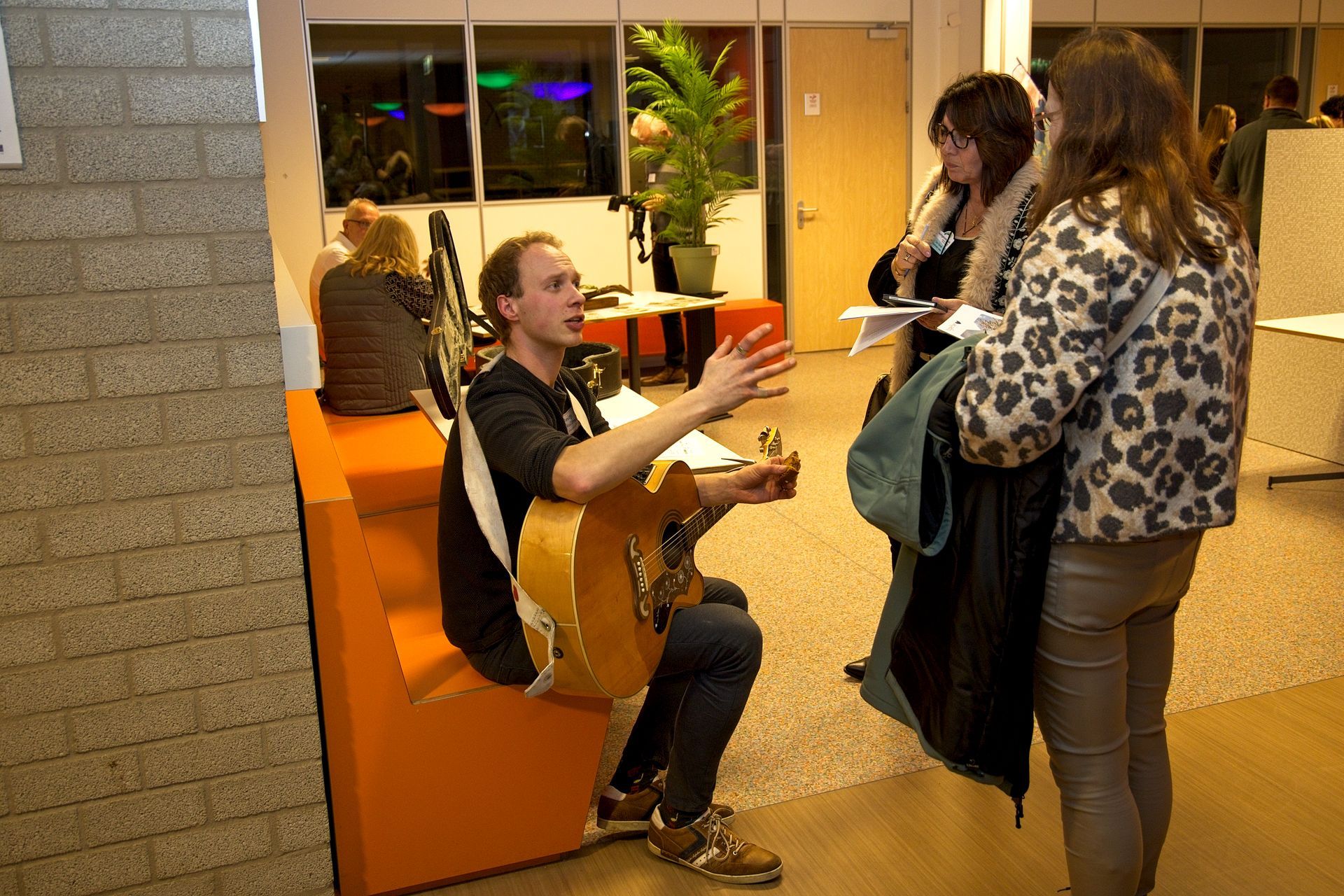Een man speelt gitaar en praat met twee vrouwen. Binnen is er een oranje bank, veel groen en een groot raam.