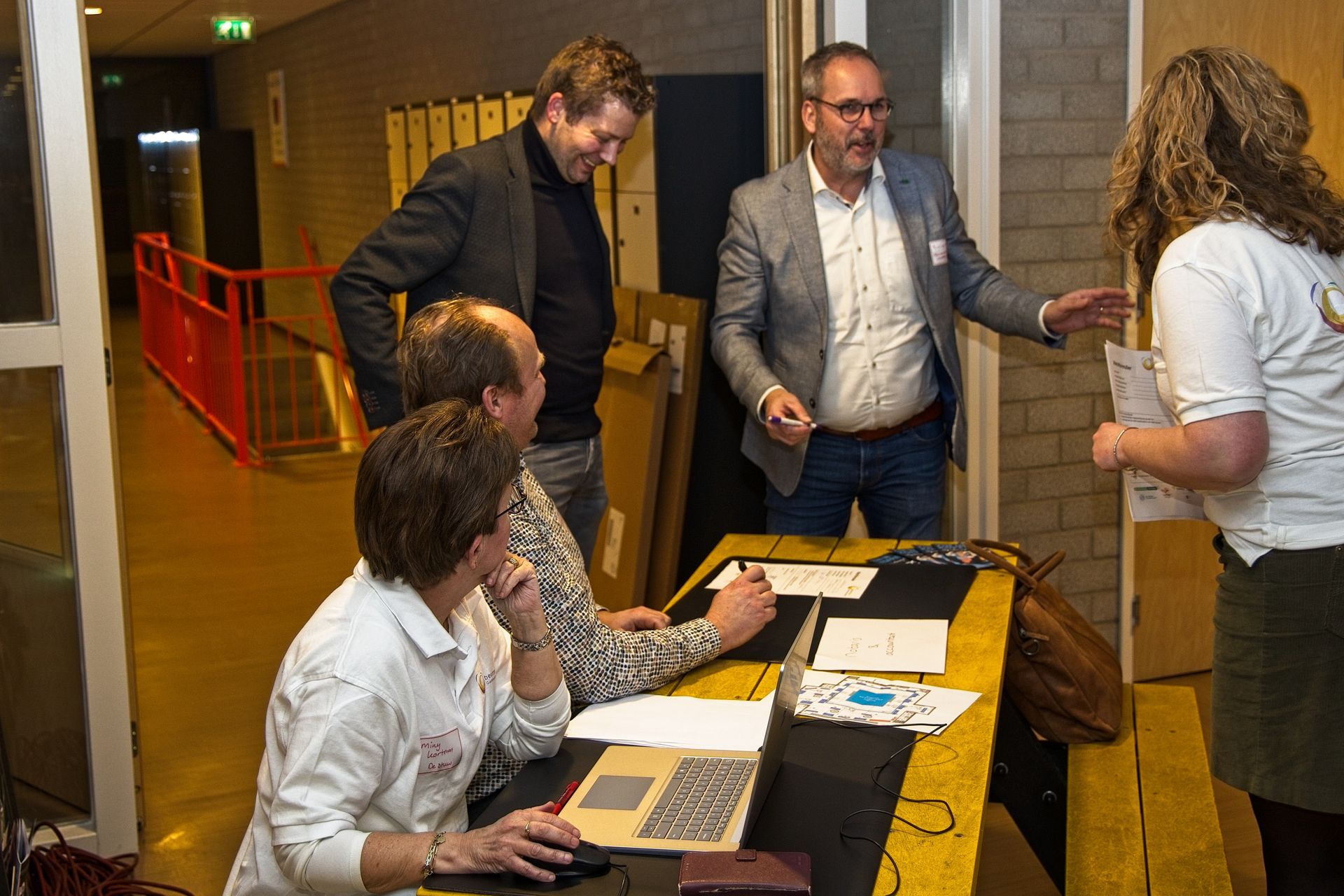 Mensen bij een inschrijfbalie, een man gebaart en controleert formulieren. Binnen in een gebouw is een rode leuning zichtbaar.