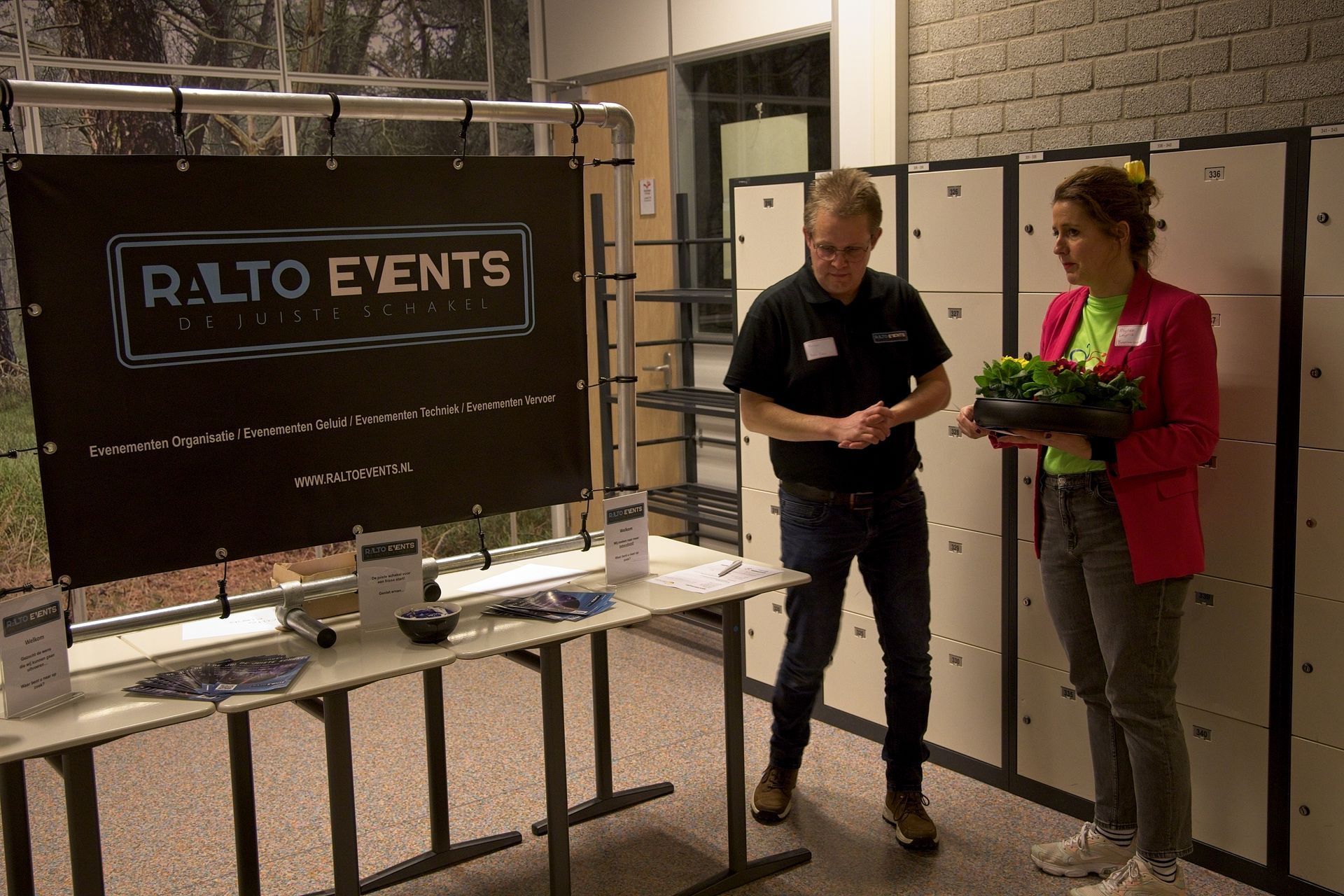 Een man en een vrouw bij een stand op een evenement met een bordje 