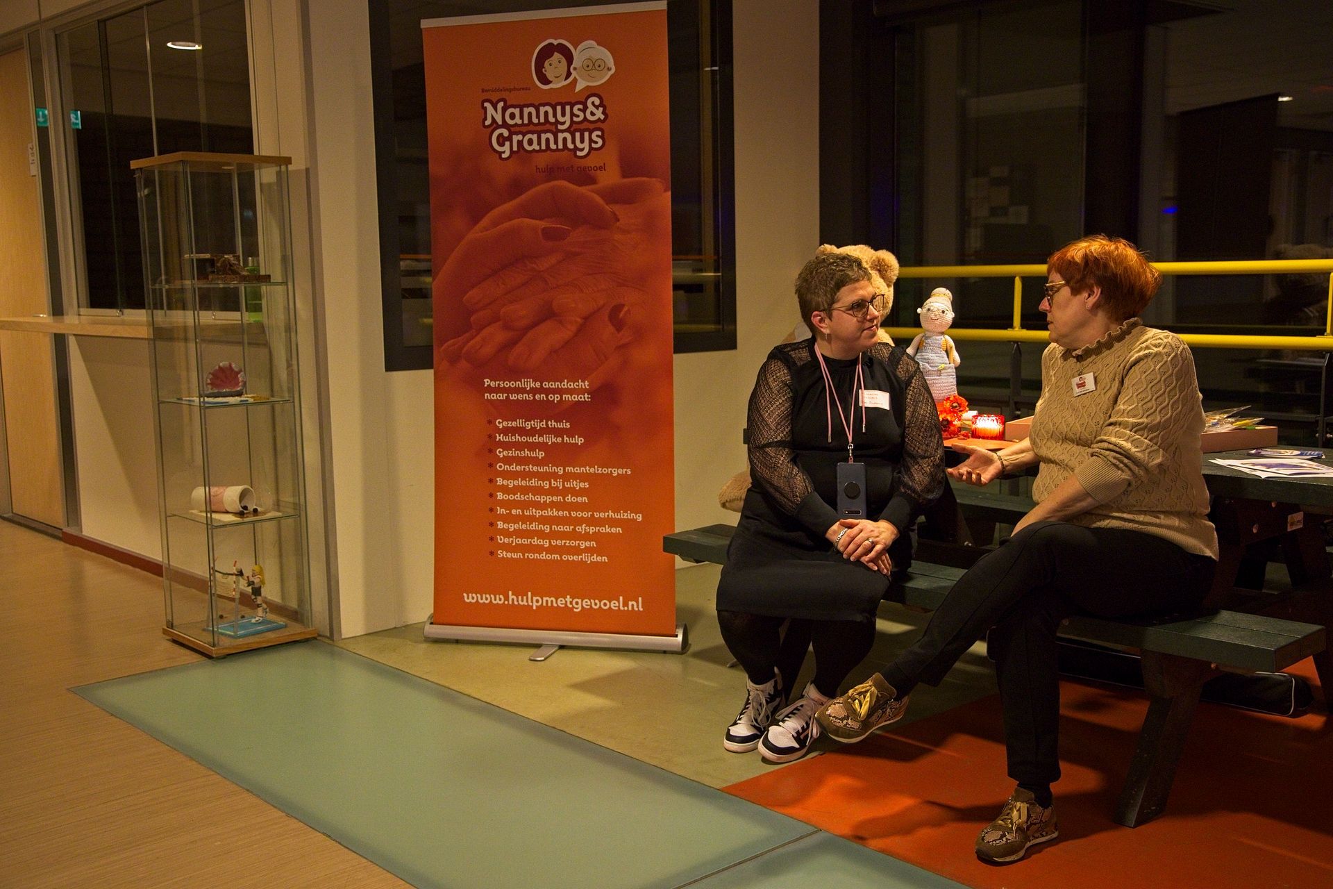 Twee vrouwen zitten op een bankje en voeren een gesprek. In de buurt, in een binnenruimte, staan ​​een oranje spandoek en een vitrine.