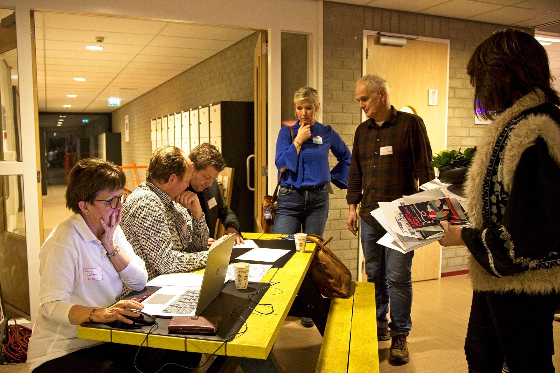 Mensen zitten aan een tafel met laptops. Anderen kijken toe, met papieren in hun handen, in een felverlichte gang.