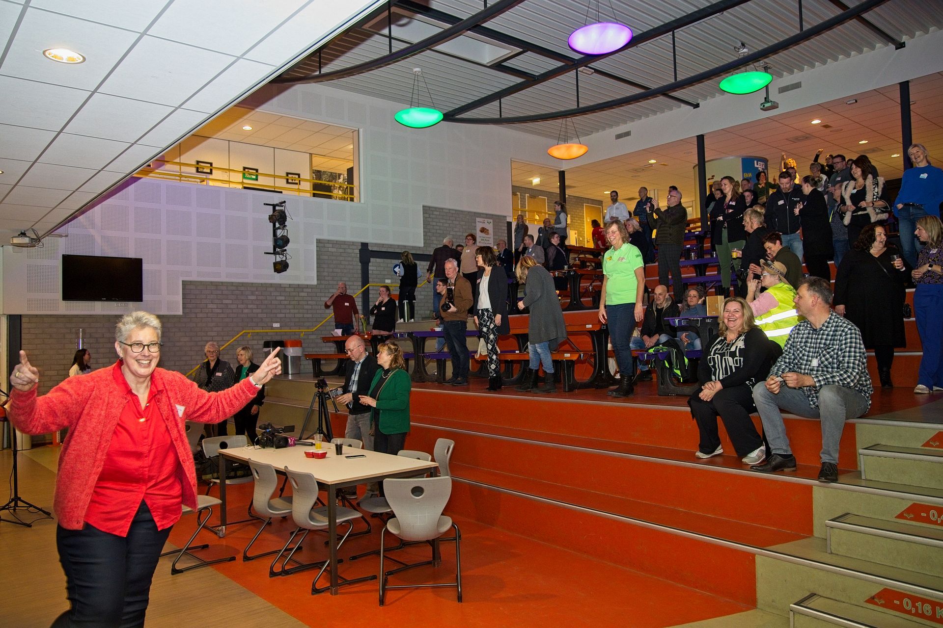Vrouw steekt haar duim omhoog tijdens een evenement; mensen zitten op een tribune. Rood, oranje, blauw, groen. Interieur.