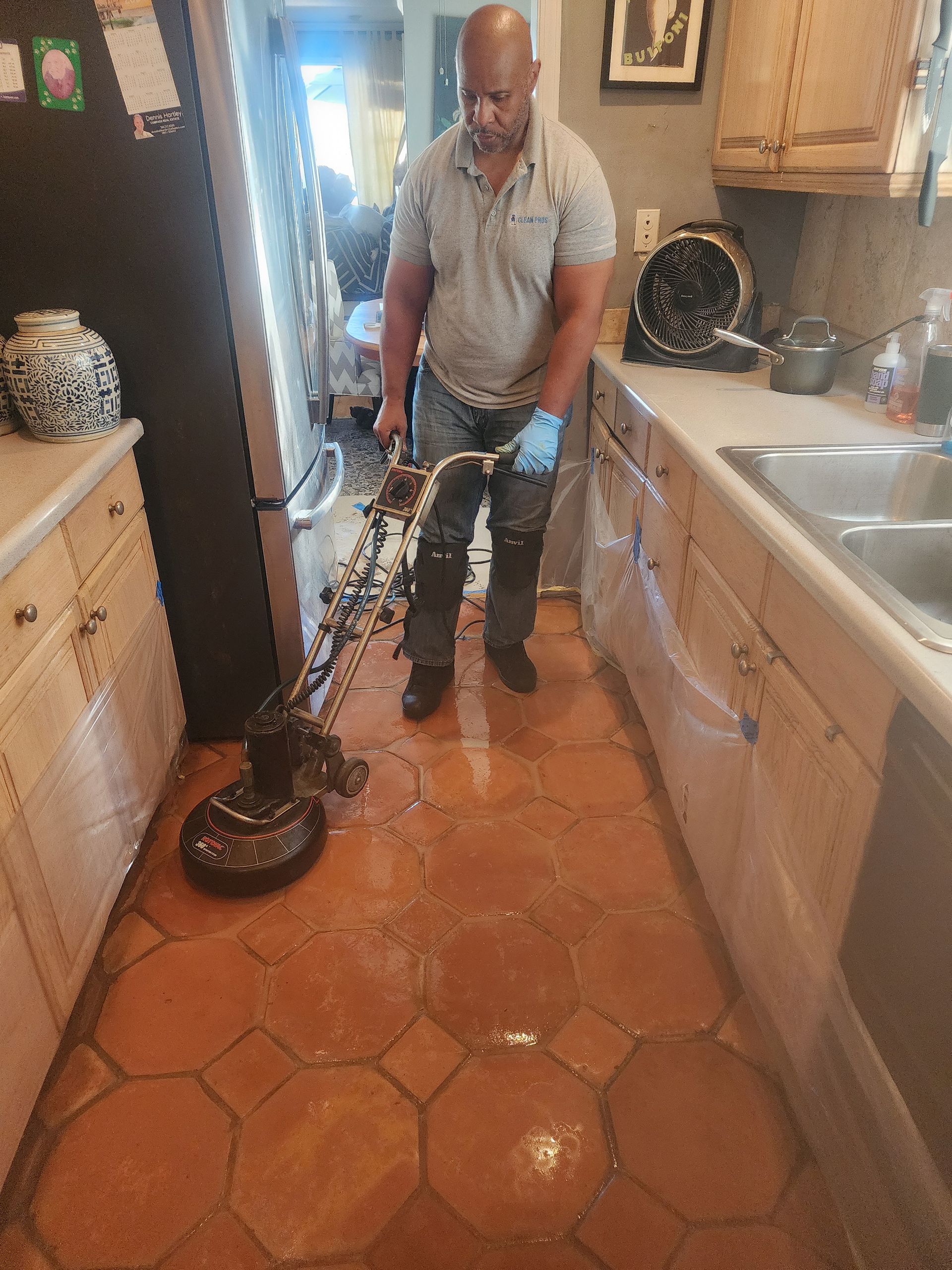 Man vacuuming a tiled kitchen floor with a canister vacuum, beside a sink and cabinets.