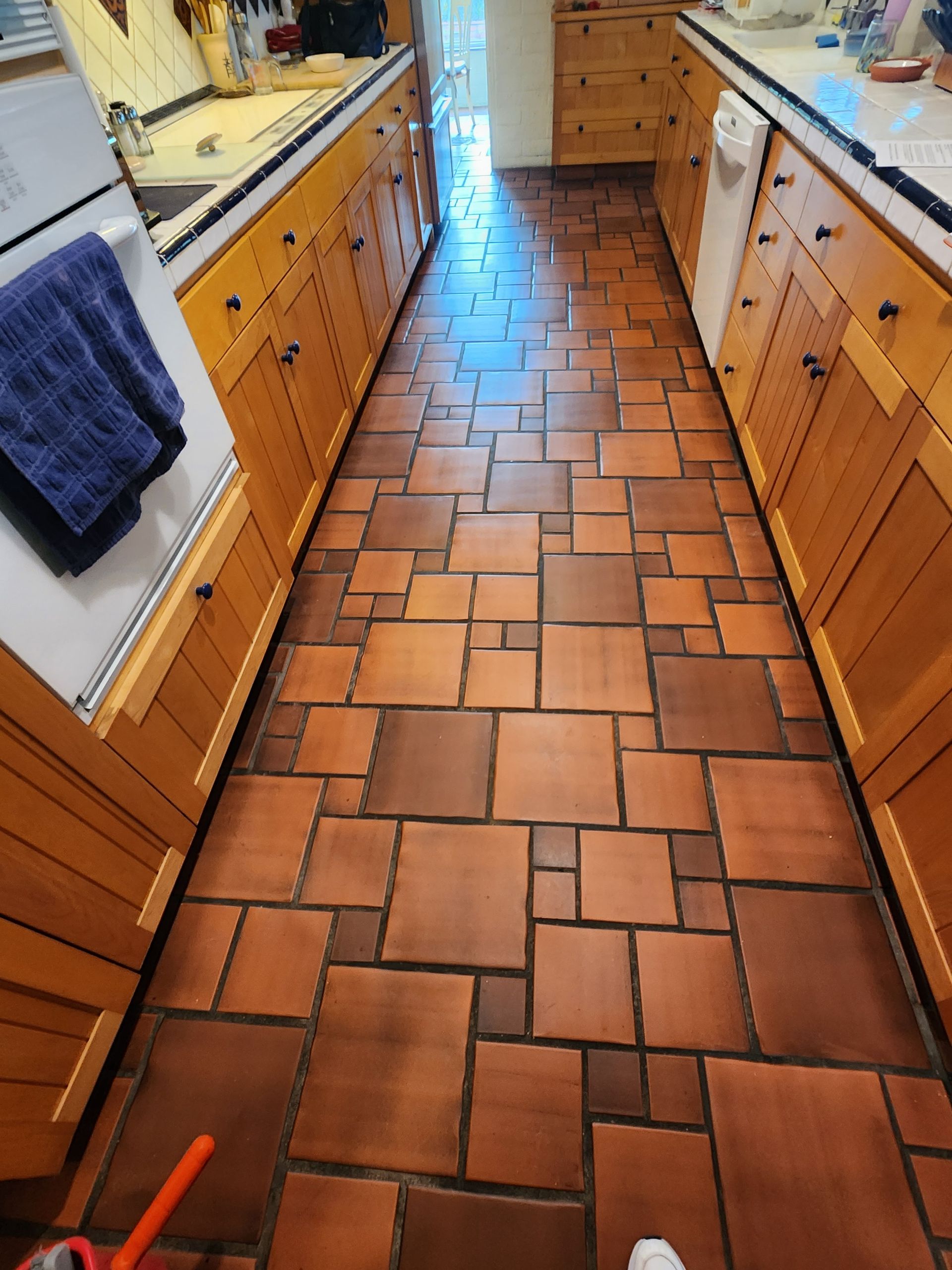 Narrow kitchen with terracotta tile floor, wood cabinets, and a towel hanging on the left side.