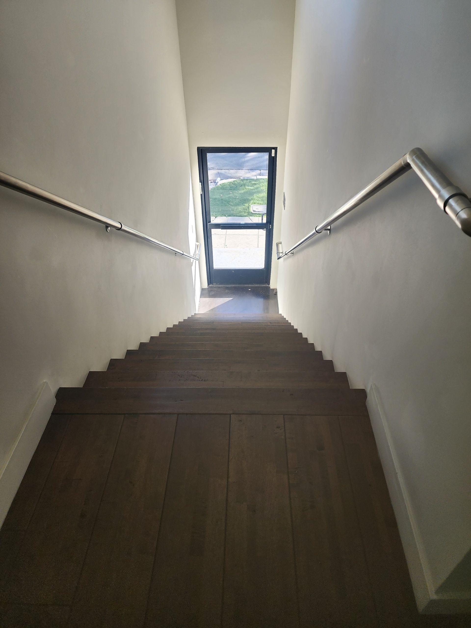 Dim stairwell with white walls, wooden steps, handrails, and a bright doorway at the bottom