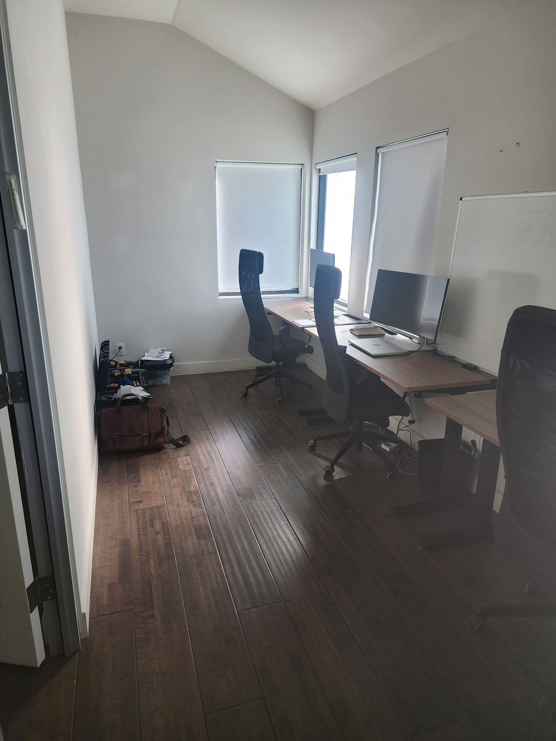 Small home office with two desks, dual monitors, office chairs, and wood floors by bright windows.