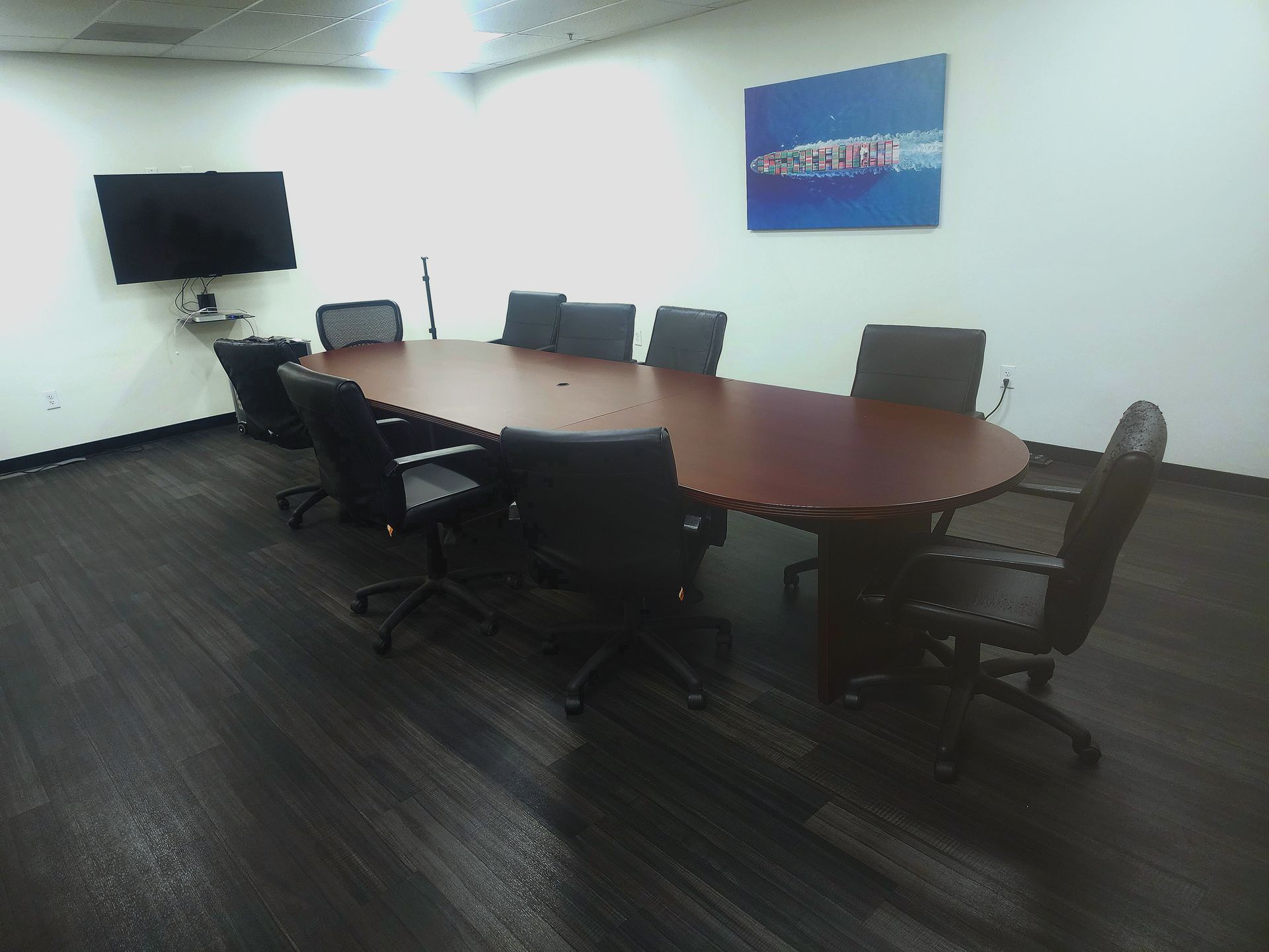 Empty conference room with oval wooden table, black chairs, wall-mounted TV, and blue wall art.