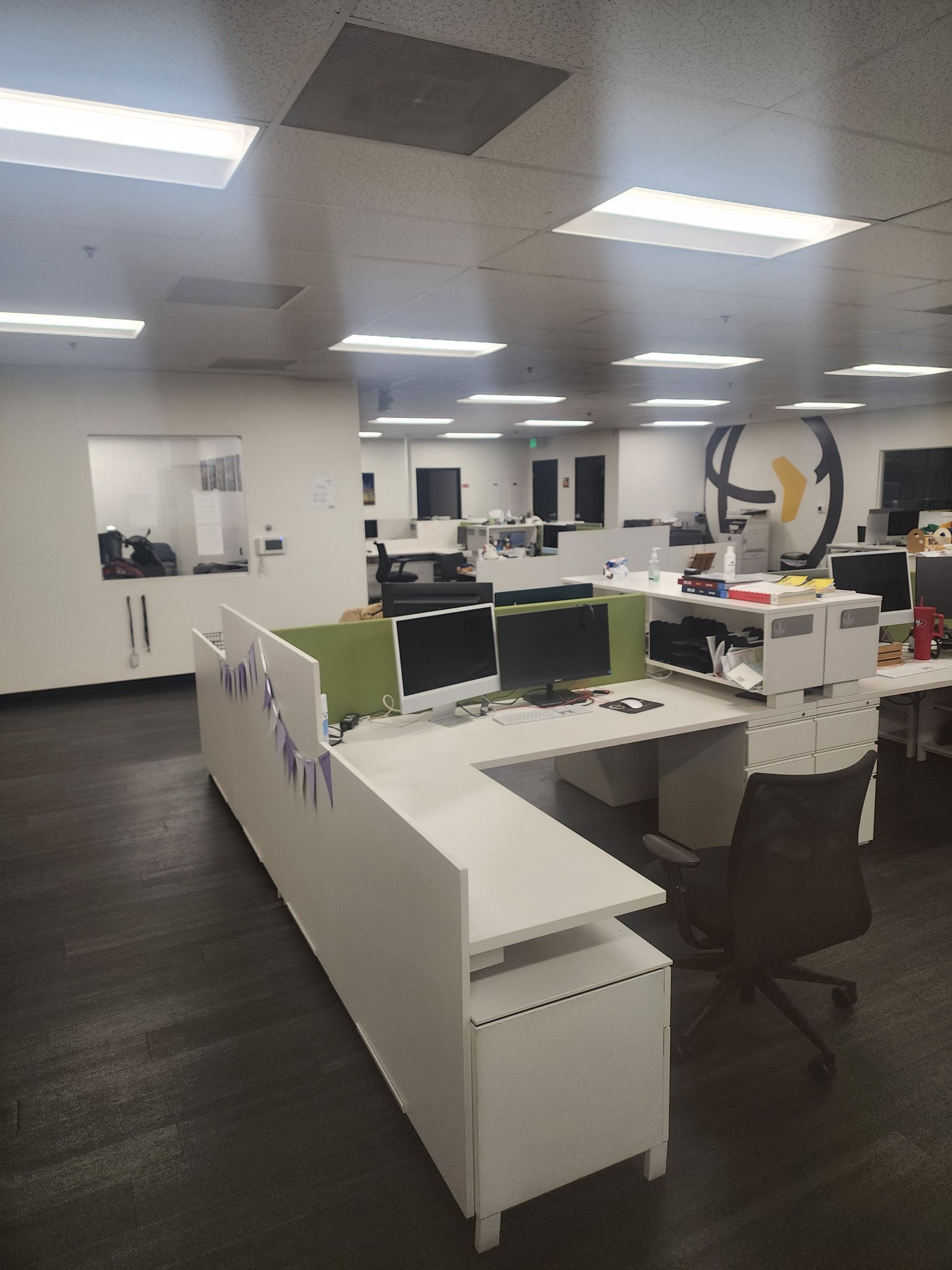 Open office workspace with desks, monitors, chairs, and bright ceiling lights