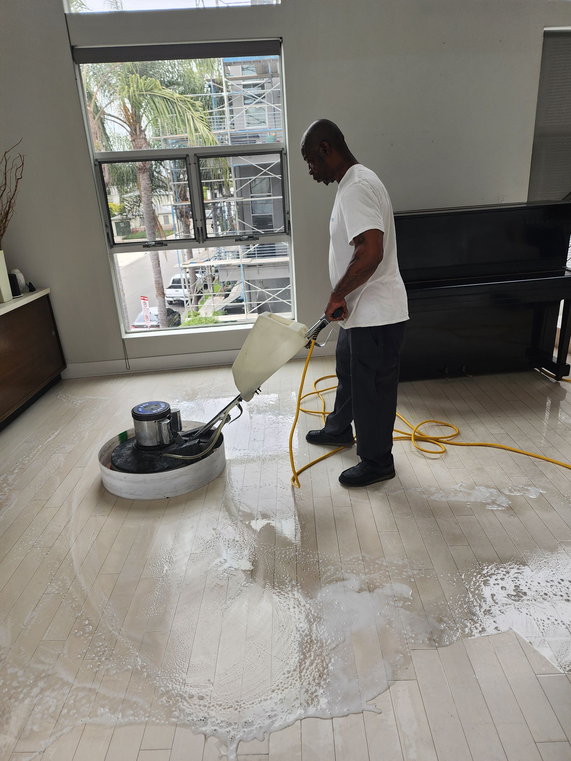 Person buffing a floor in a bright room, with cleaning solution spraying near a large window.