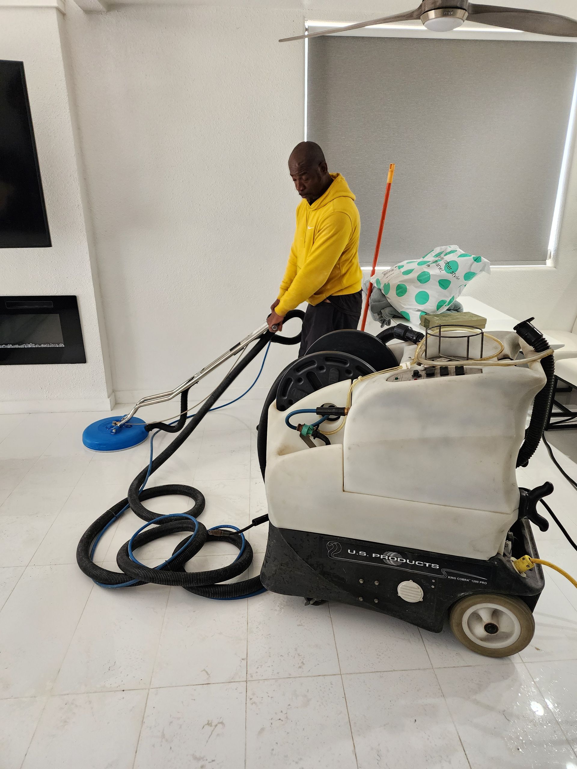 Worker using a floor-cleaning machine in a white room with a black TV stand and a mop bucket nearby.