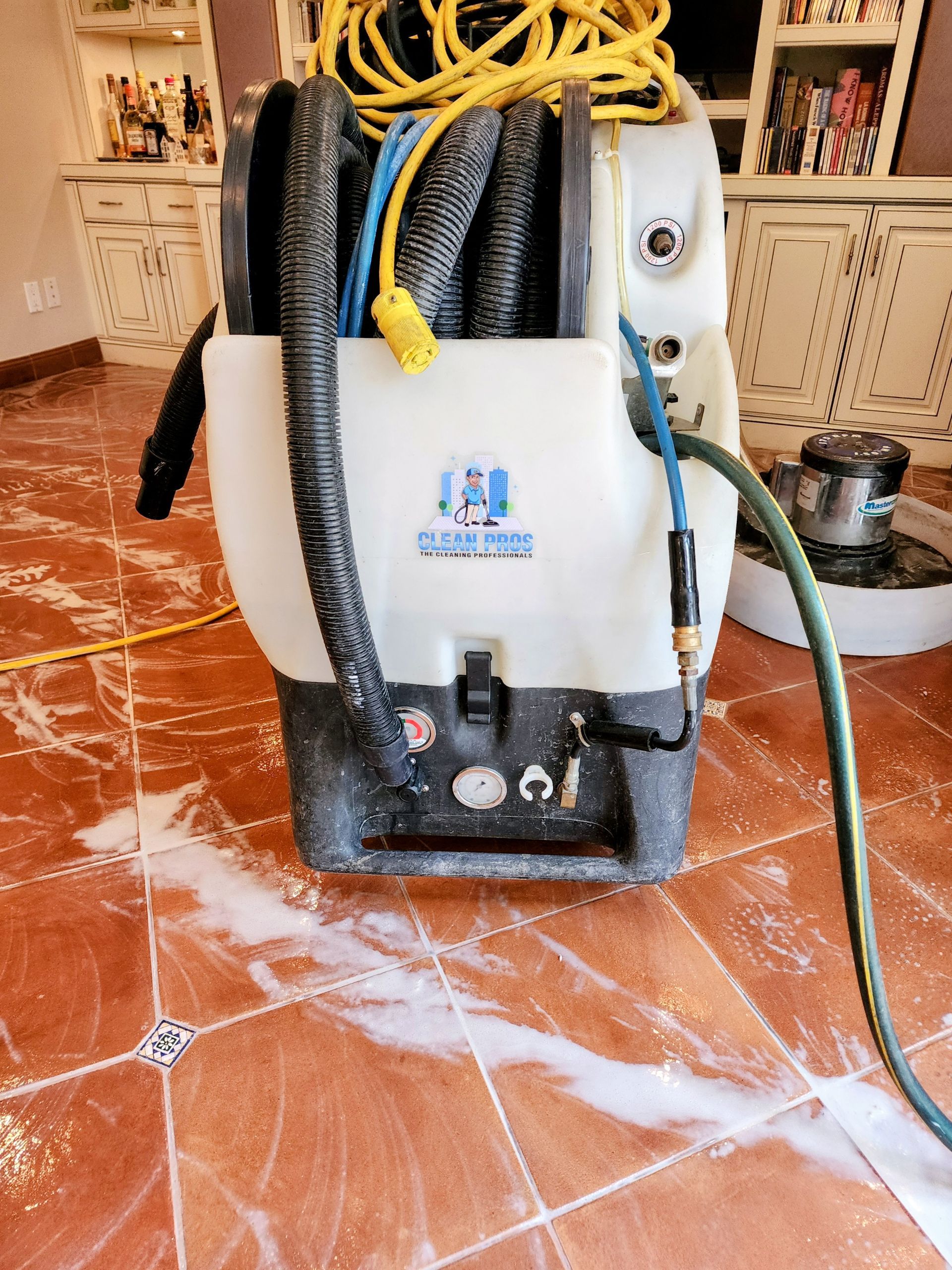 A steam cleaning machine with hoses and yellow cords on a wet, soapy tiled floor indoors