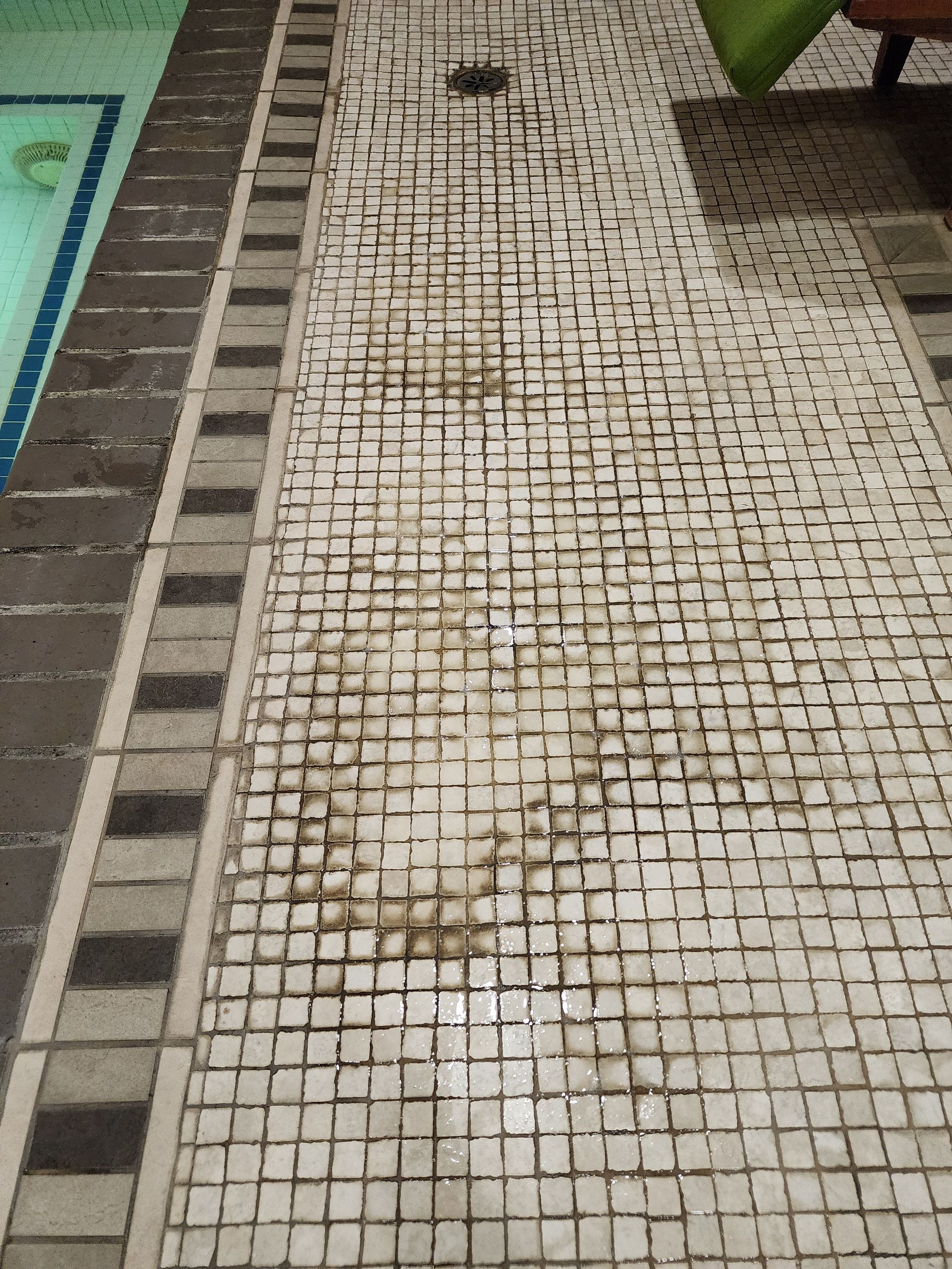 Narrow outdoor walkway with white mosaic tiles and dark border tiles beside a pool edge.