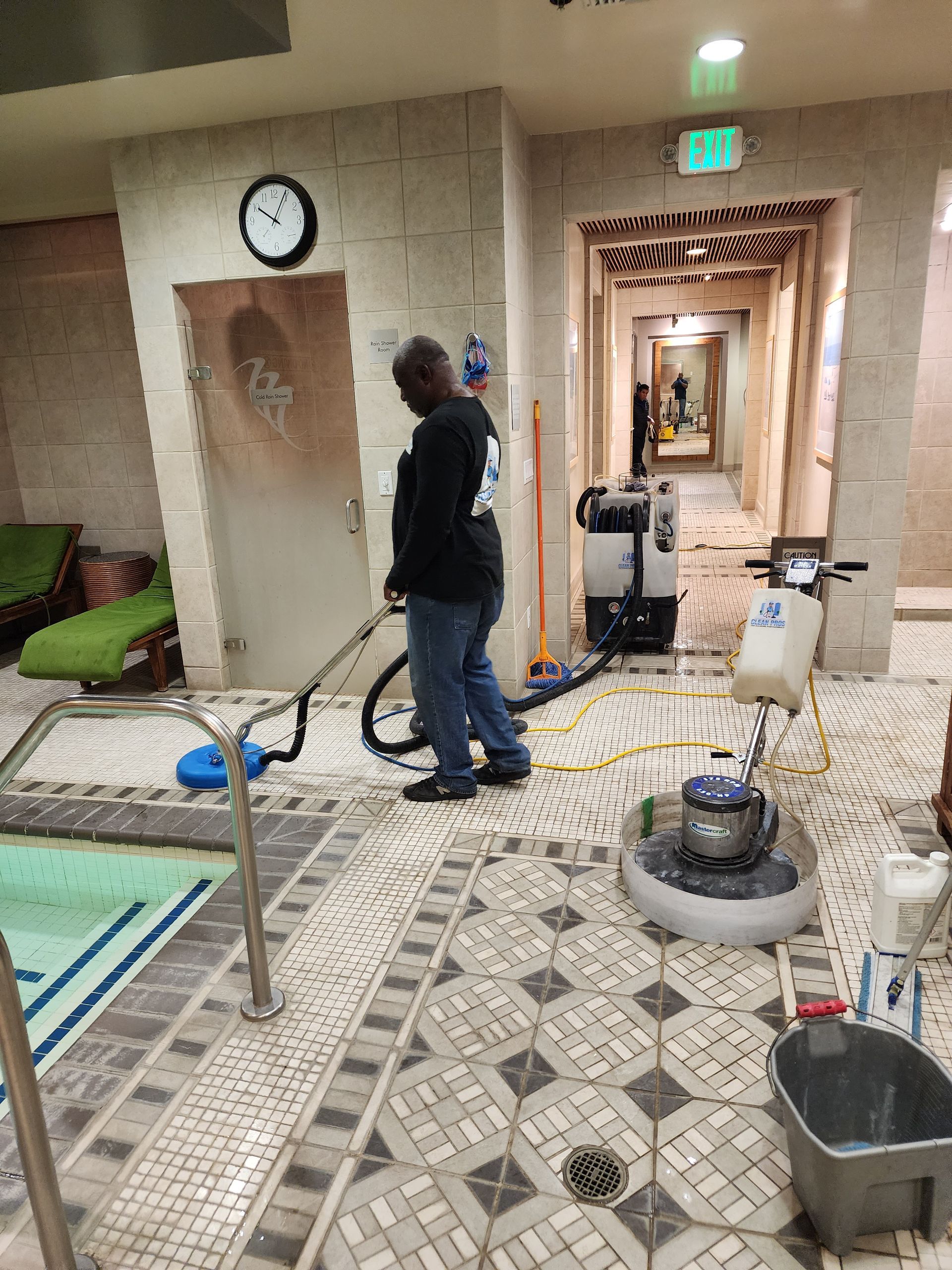 Man in a hotel hallway using a floor buffer, with cleaning tools and equipment nearby.