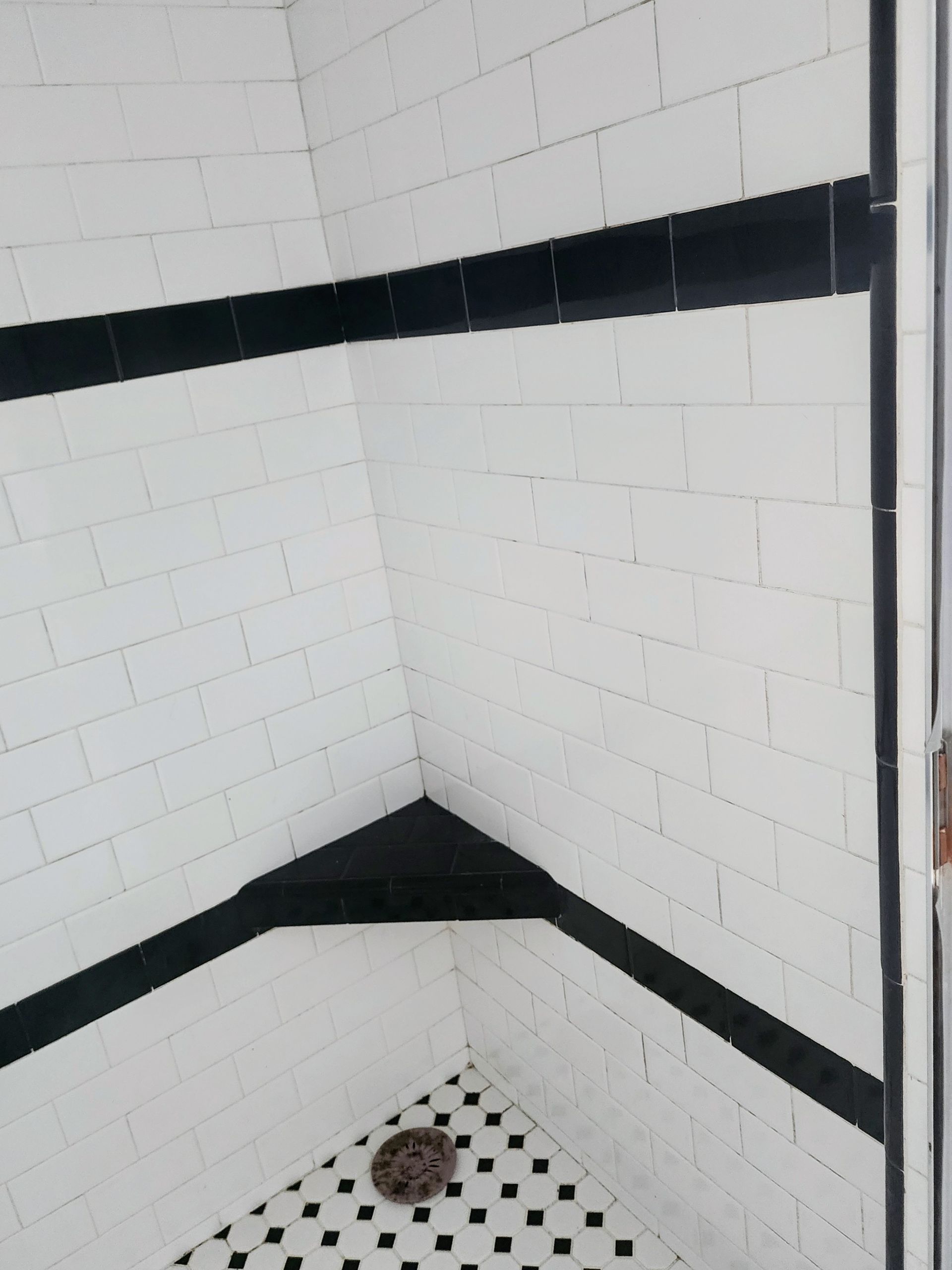 White tiled shower corner with black trim and a drain at the bottom.