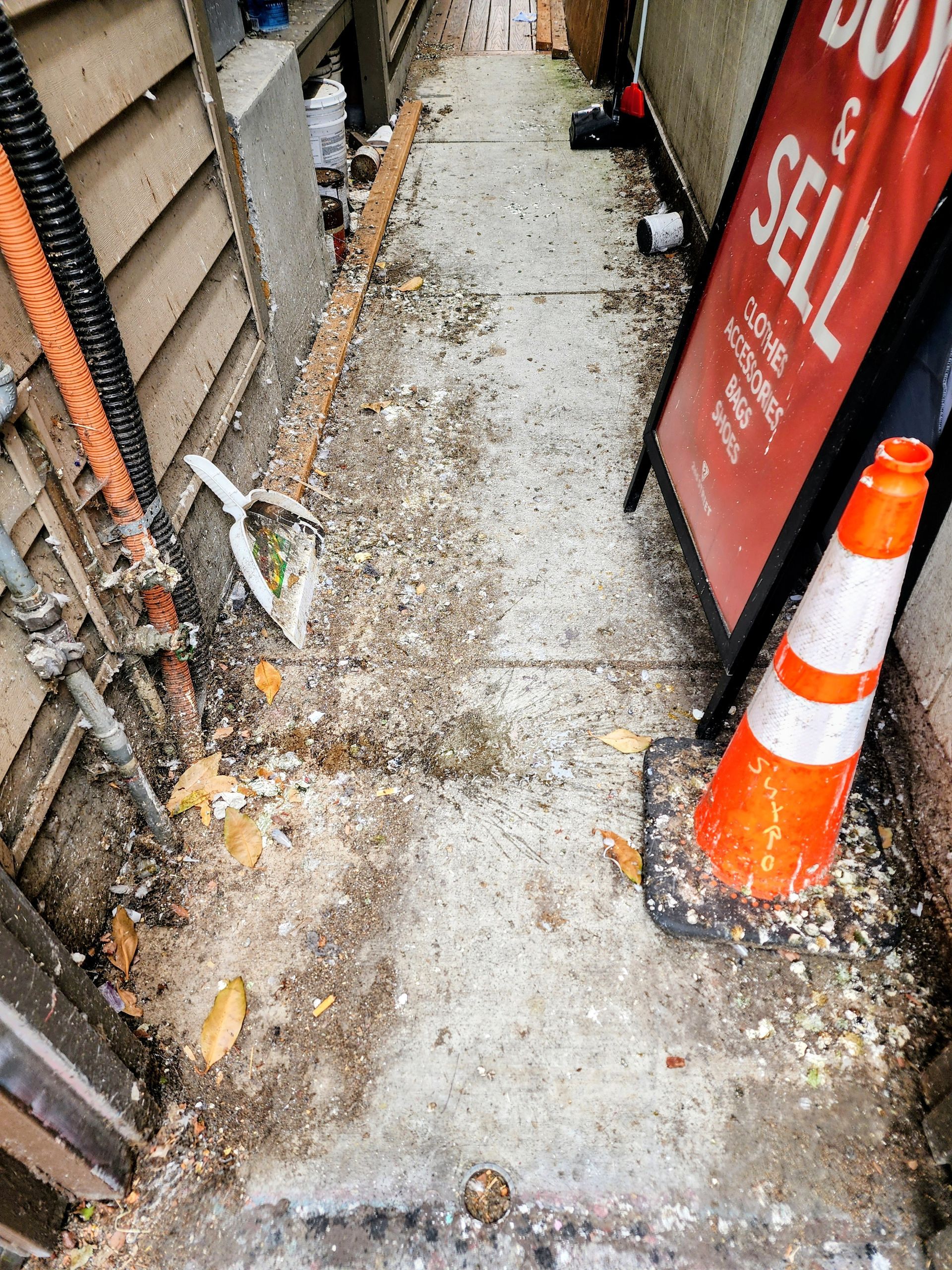 Narrow gravel alley with orange traffic cone and red sign panel beside a building.