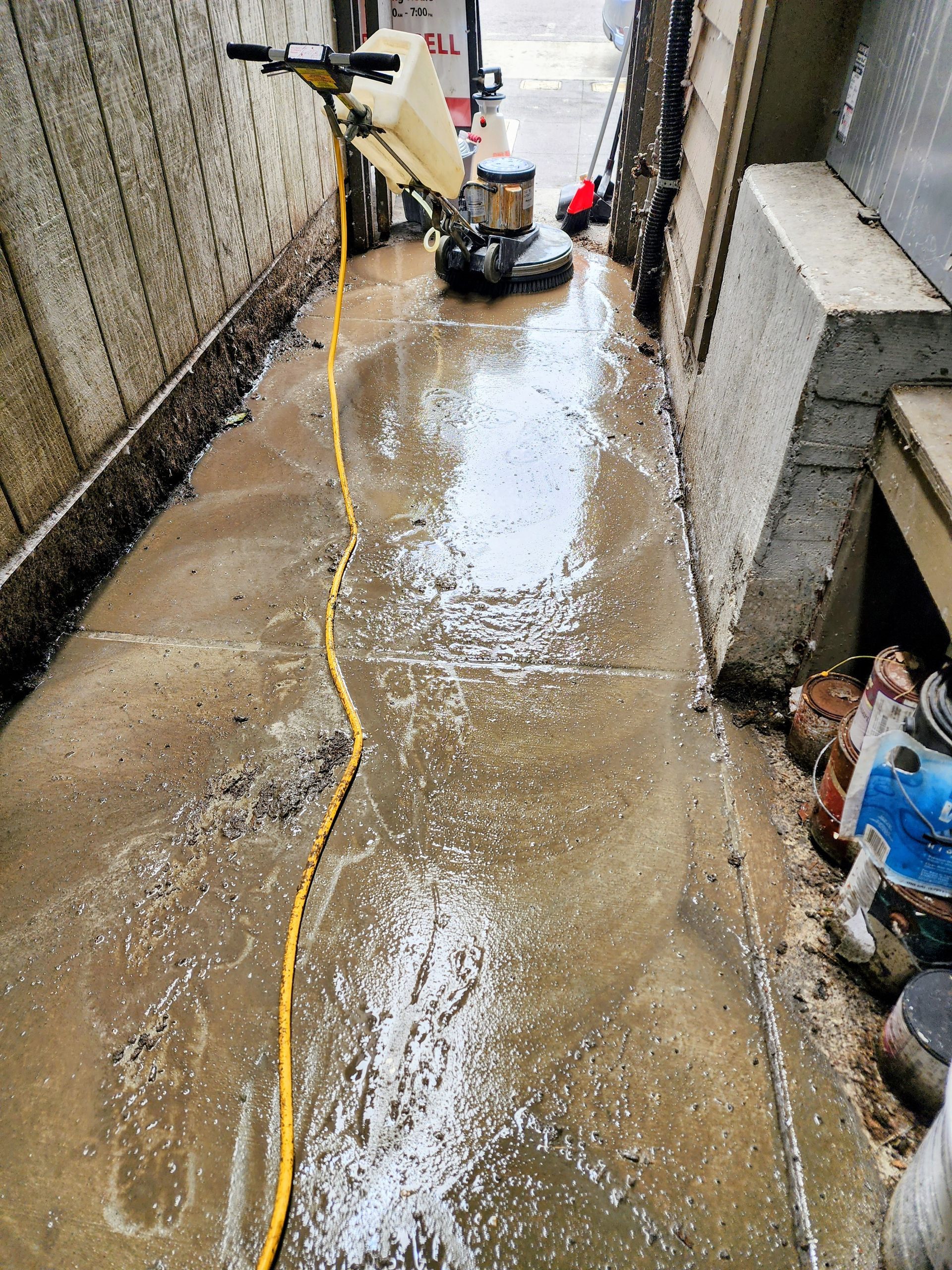 Pressure washer cleaning a narrow alley, with wet concrete and a yellow hose running down the path.