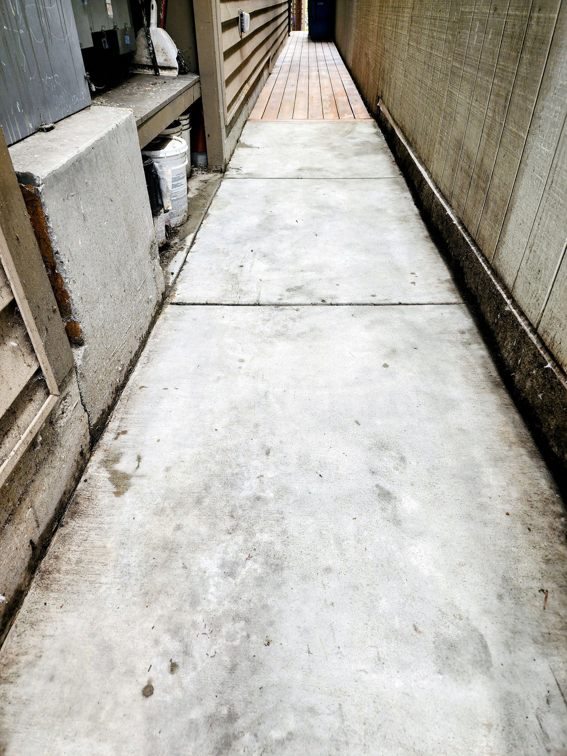 Narrow concrete walkway between buildings, leading to a wooden bridge ahead.