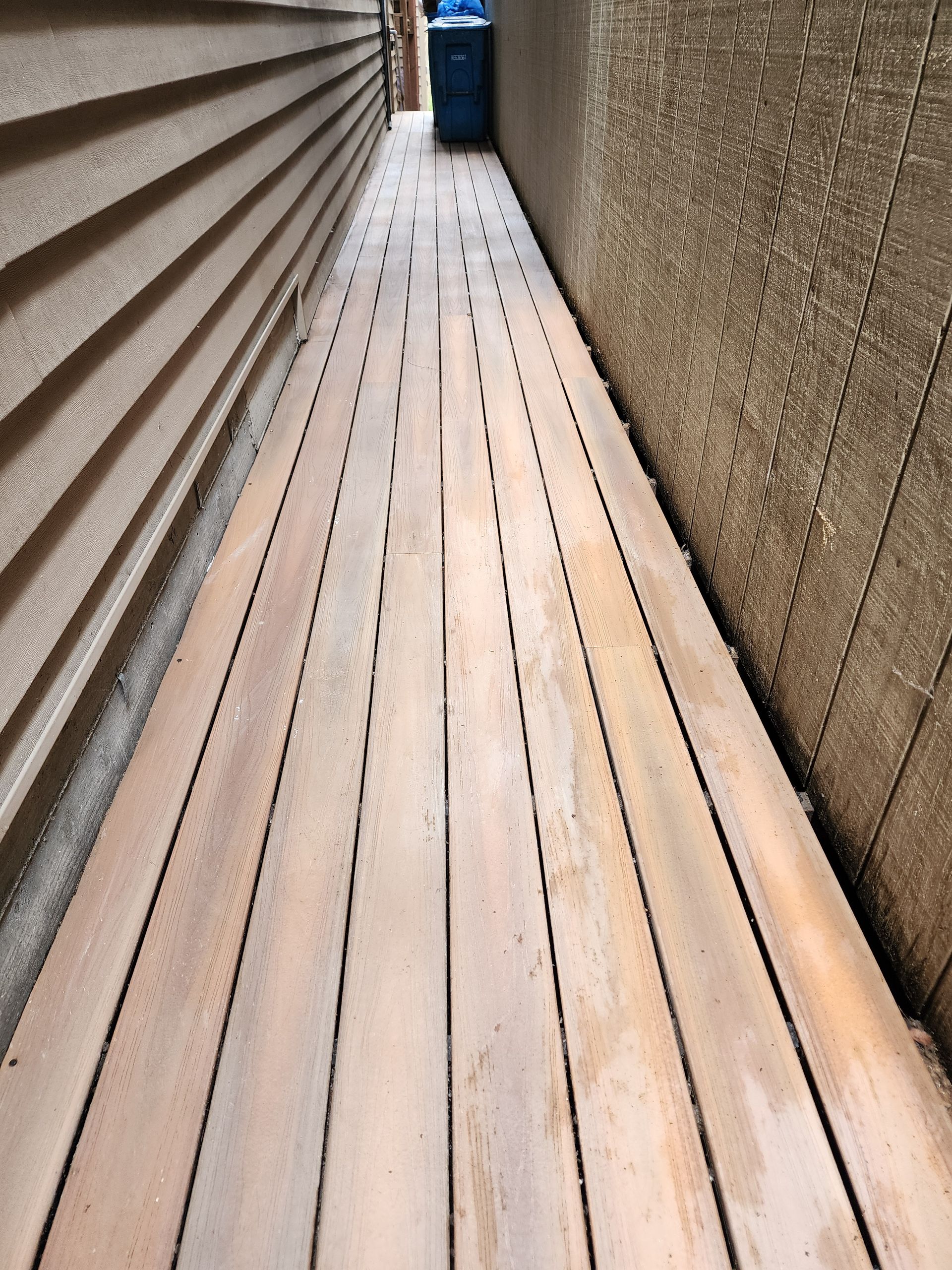 Narrow outdoor walkway with wooden planks between two textured walls, leading to a blue bin in the distance