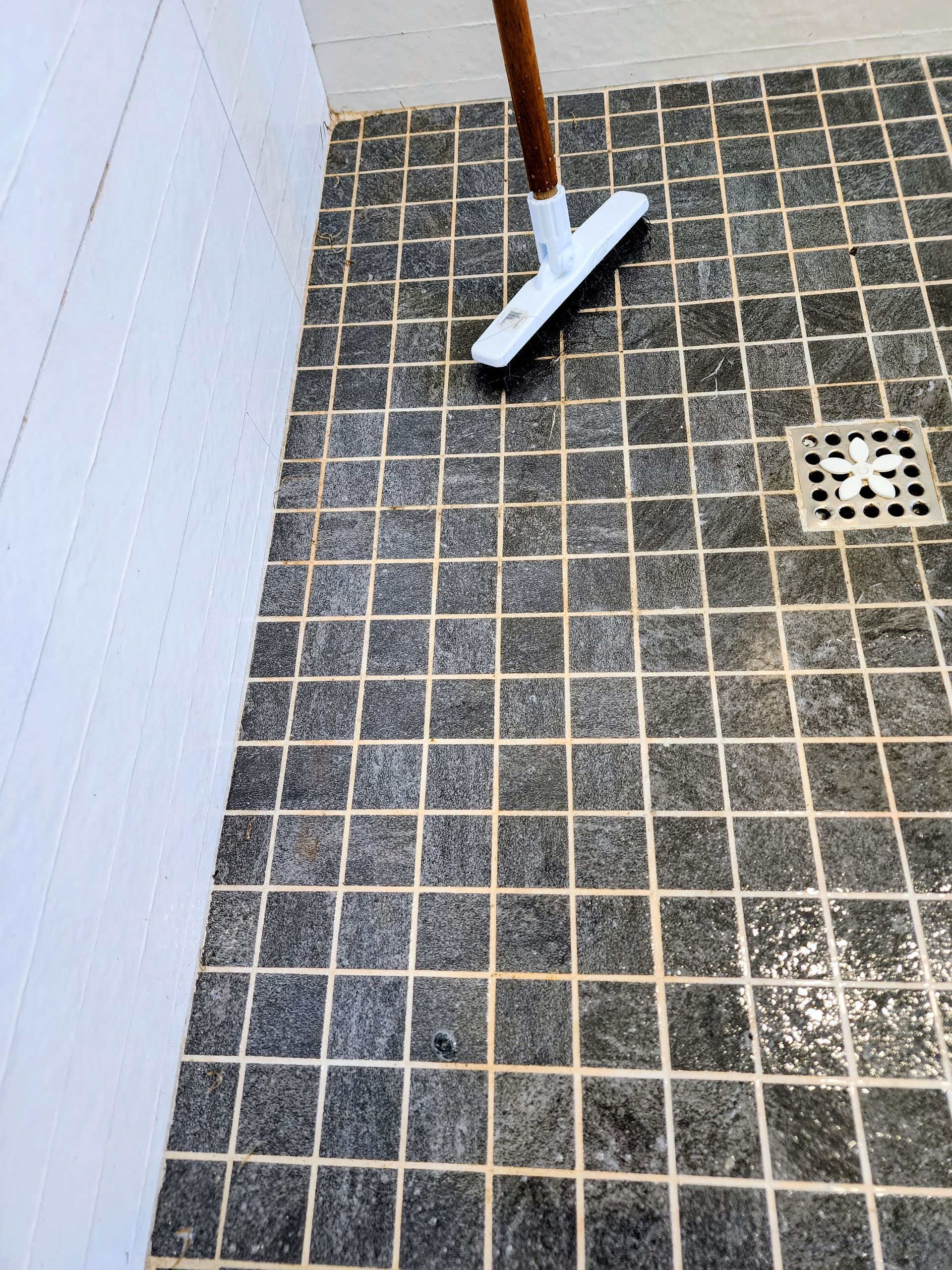 White broom on dark tiled bathroom floor near a floor drain and white wall