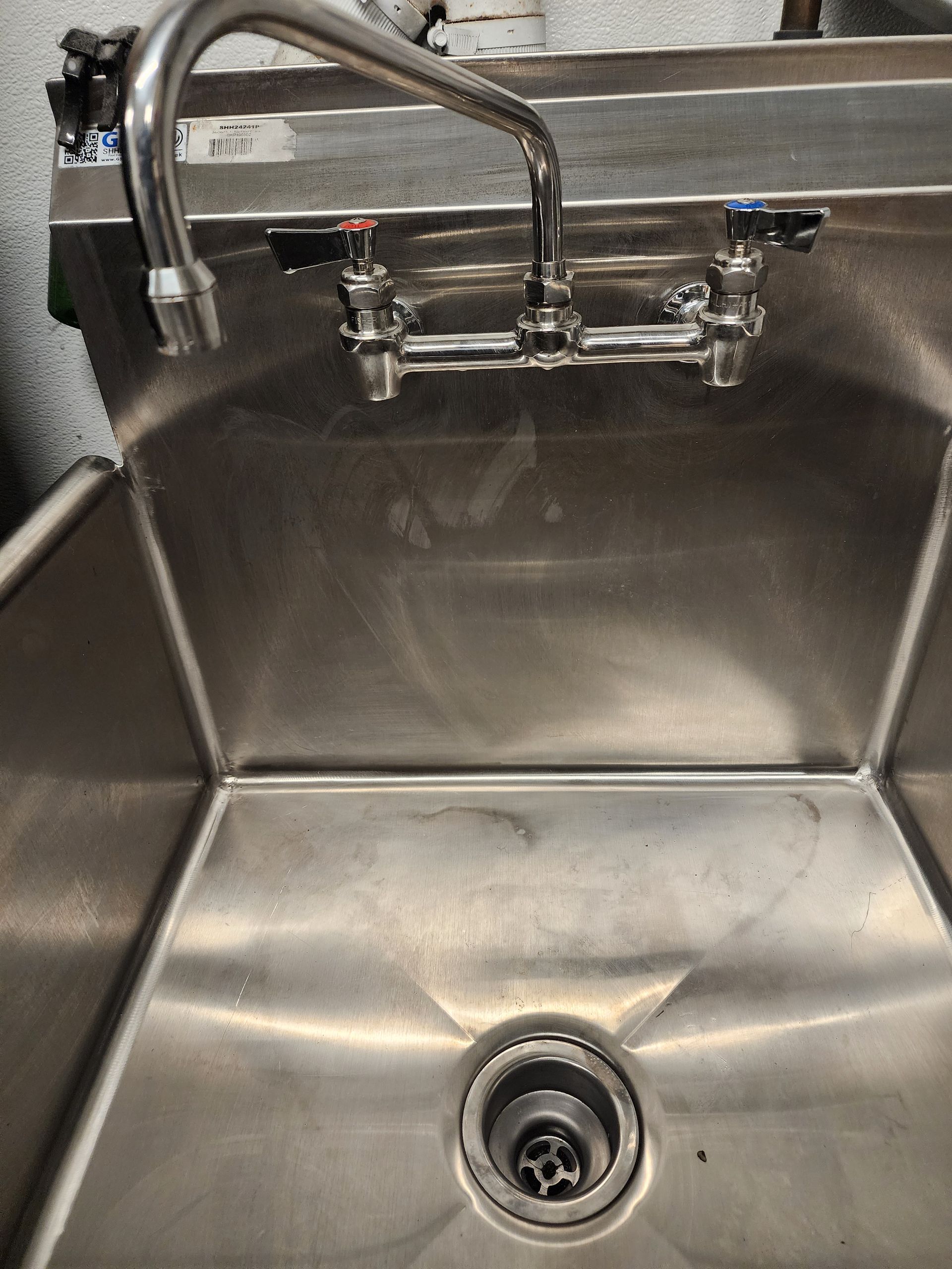 Commercial stainless-steel sink with dual faucets and a drain in the basin.