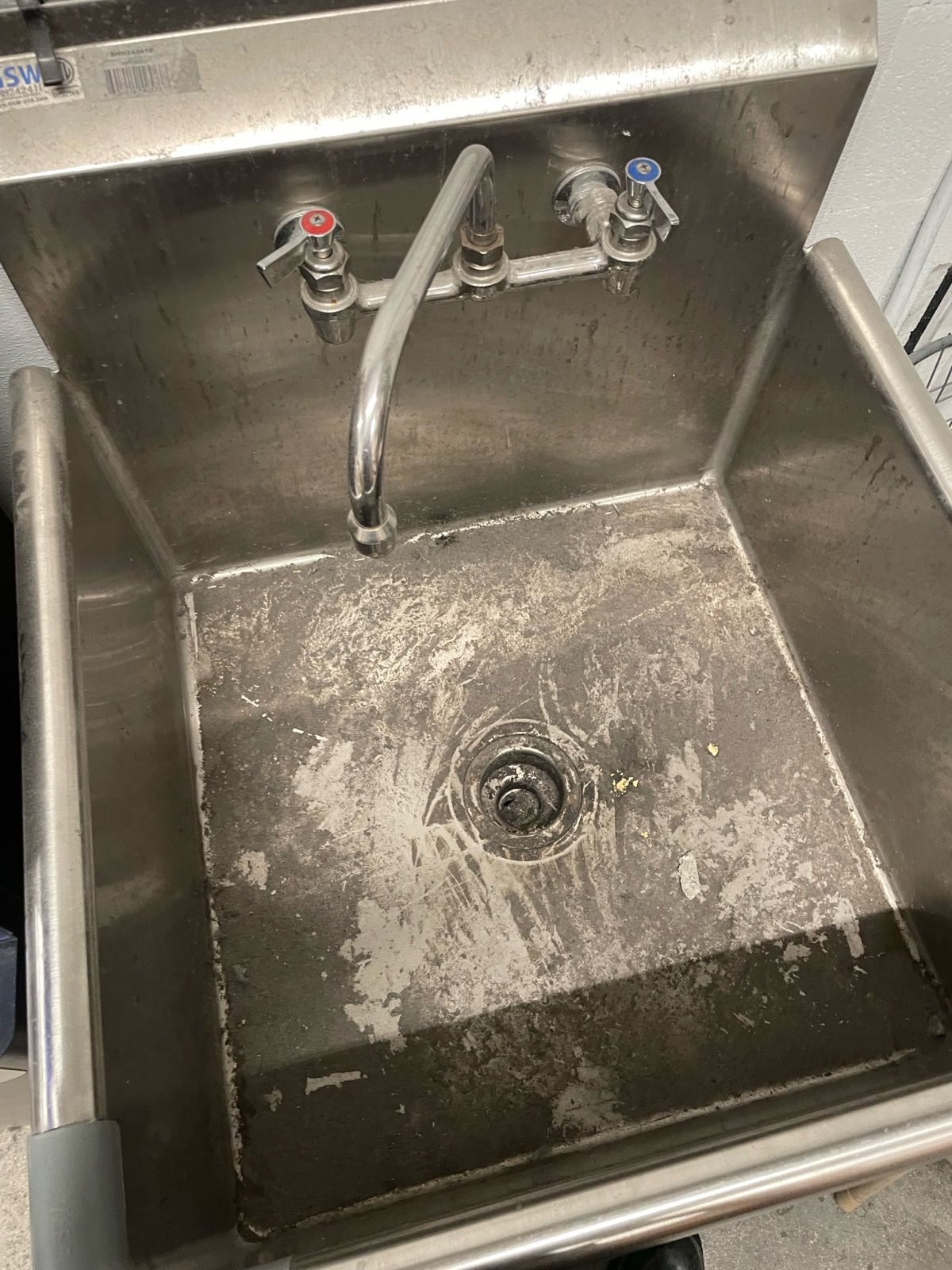 Stainless steel utility sink with two faucets and a central drain, showing water stains and wear.