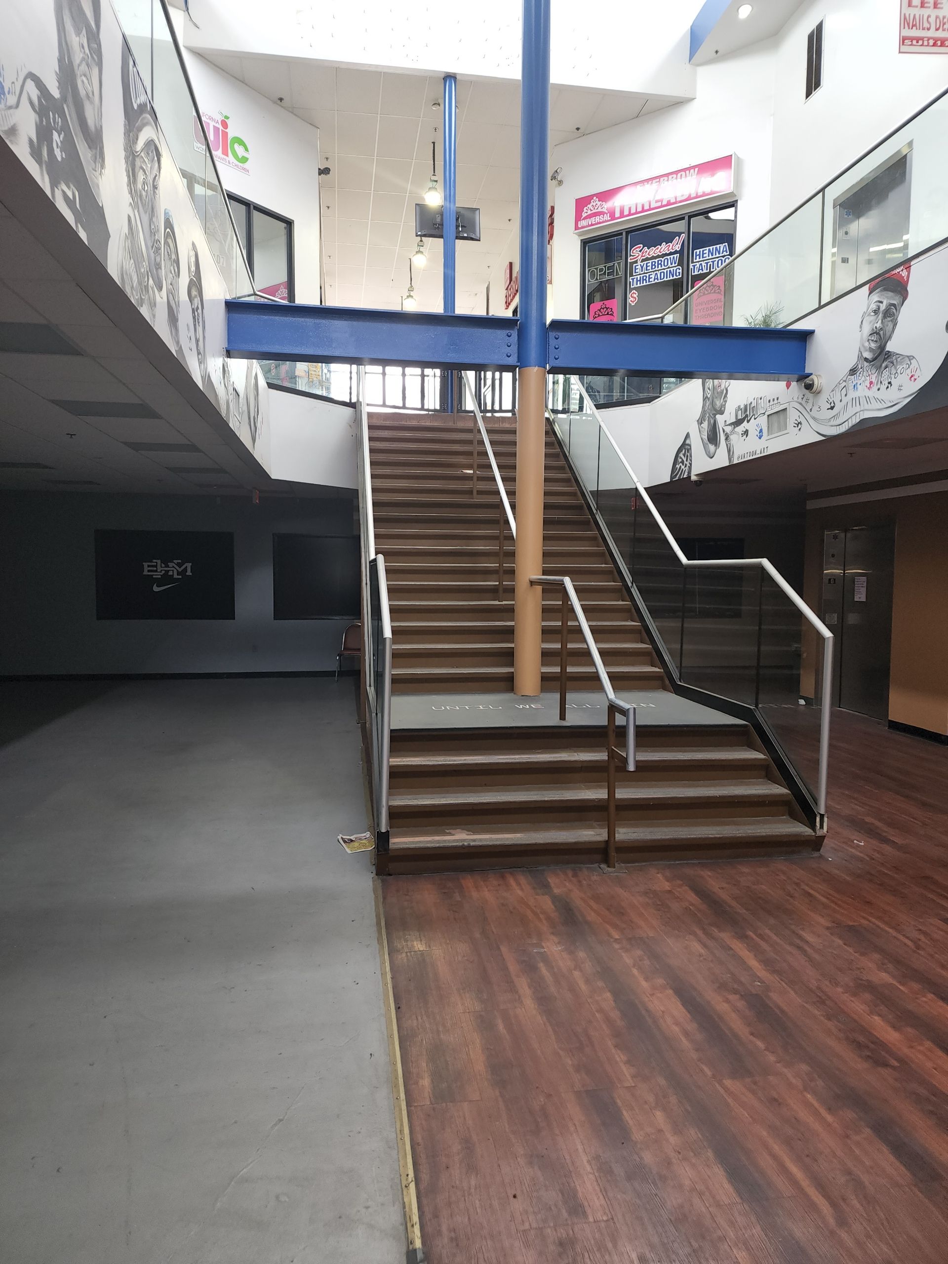 Indoor atrium with a central staircase, blue railings, and wood flooring beneath a skylit ceiling.