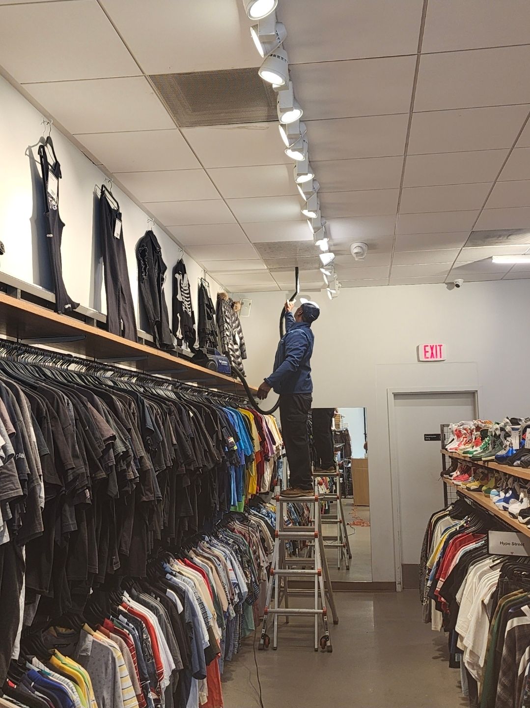 Person reaching up to adjust a clothing store ceiling light among hanging shirts and jackets