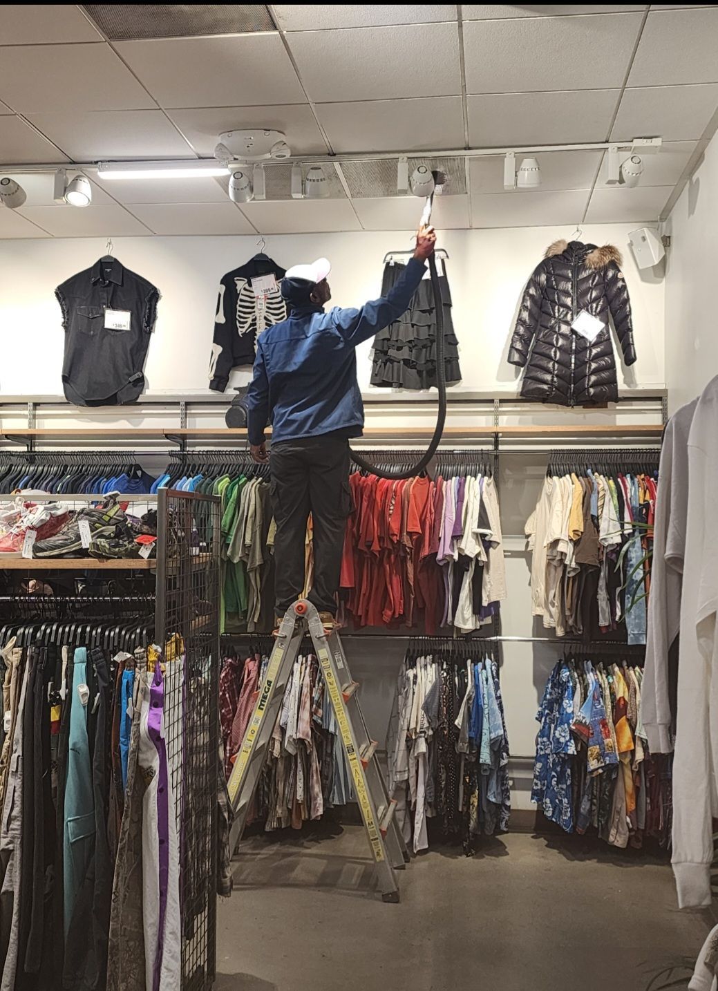 Person reaching for a shirt on a high shelf in a clothing store, surrounded by hanging clothes.