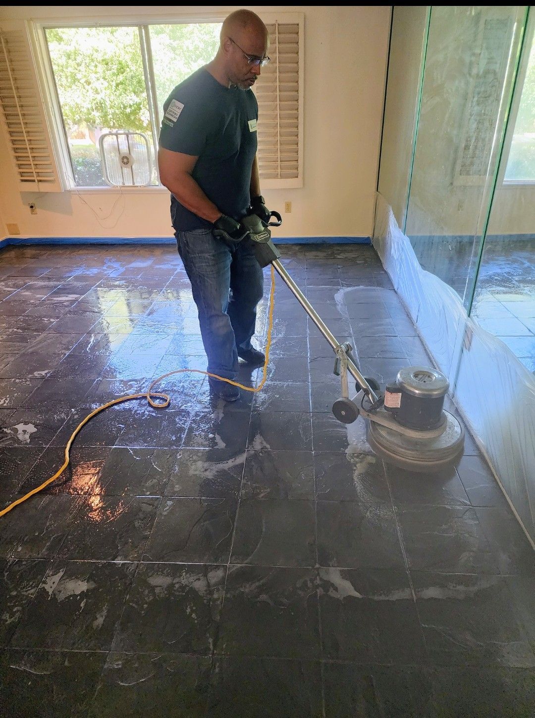Worker polishing a dark tile floor with a floor buffer in a sunlit room