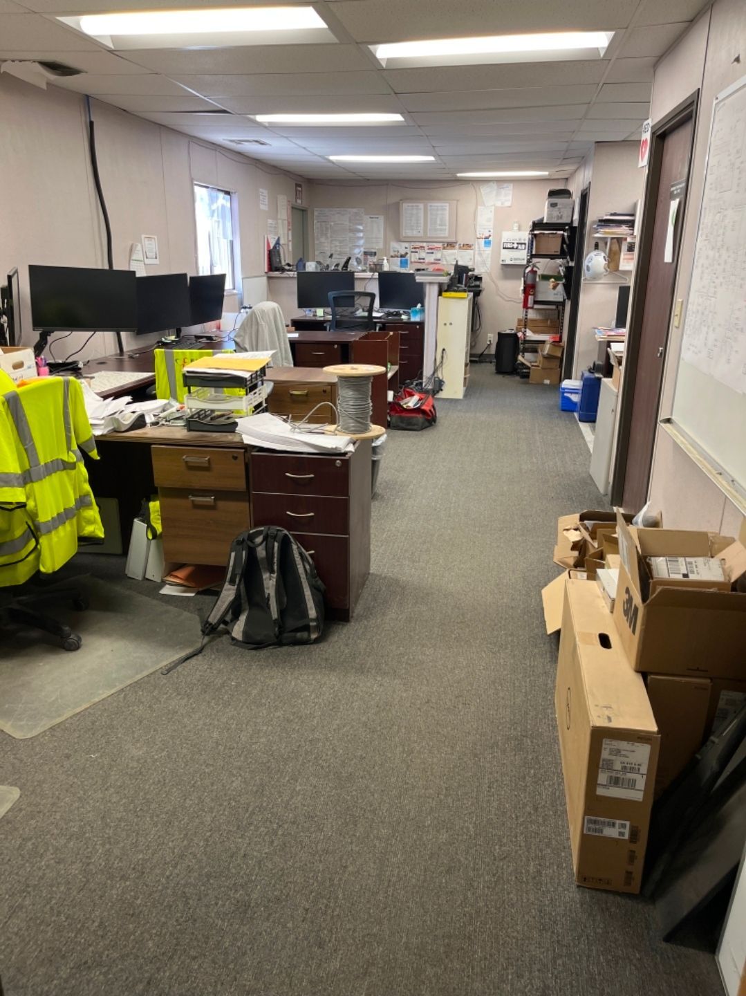 Cluttered office hallway with desks, boxes, shelves, and a bright yellow safety vest draped on a chair.