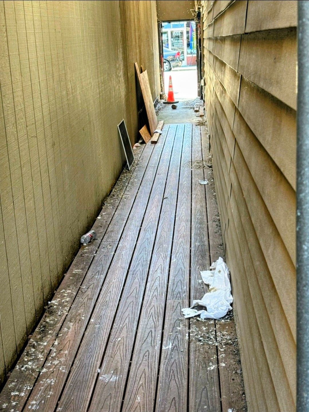 Narrow wooden walkway between tall walls, with scattered debris and a bright opening at the end.