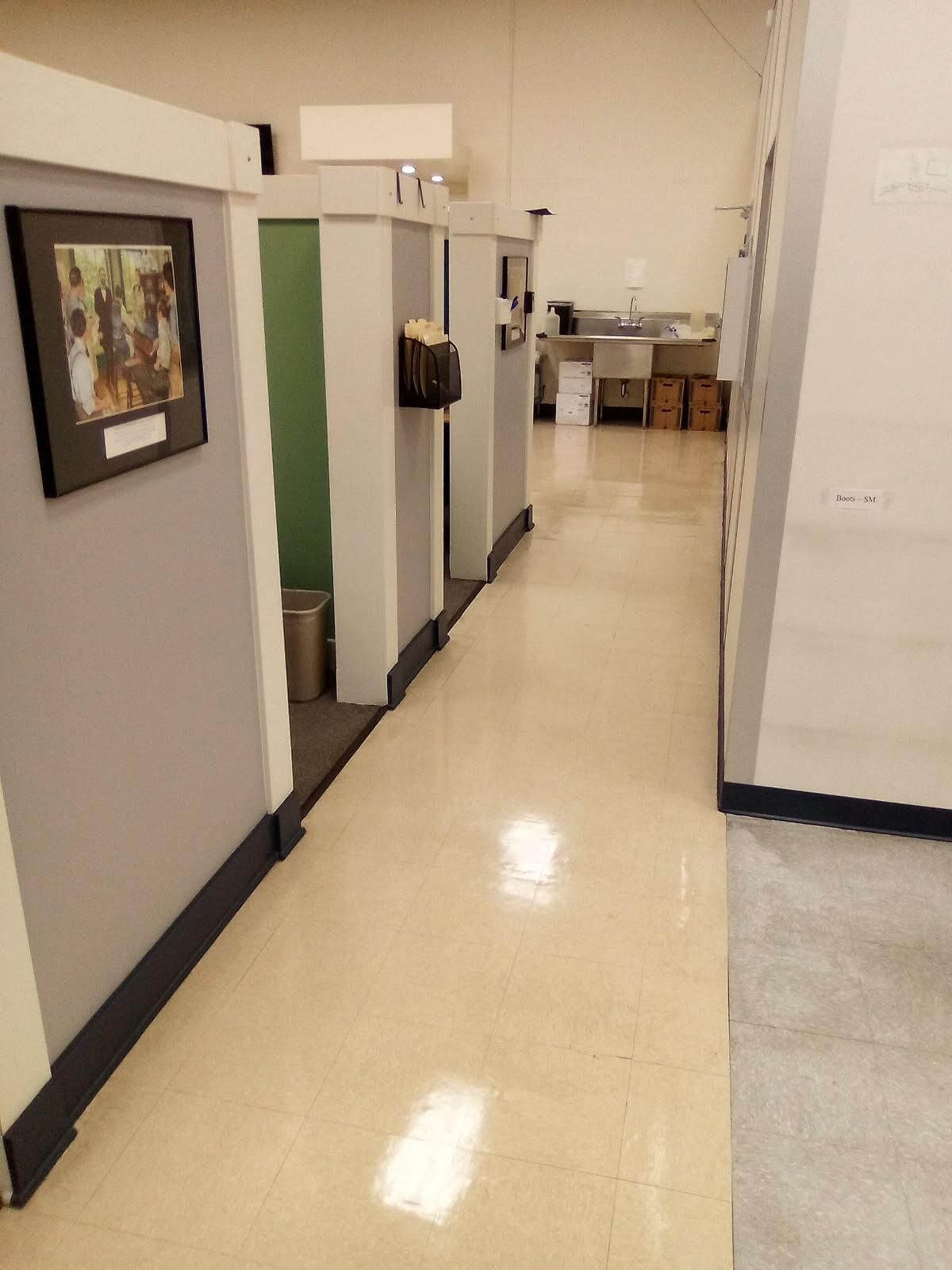 Empty hallway with beige tile floor, gray walls, and a few framed pictures and office chairs in the distance
