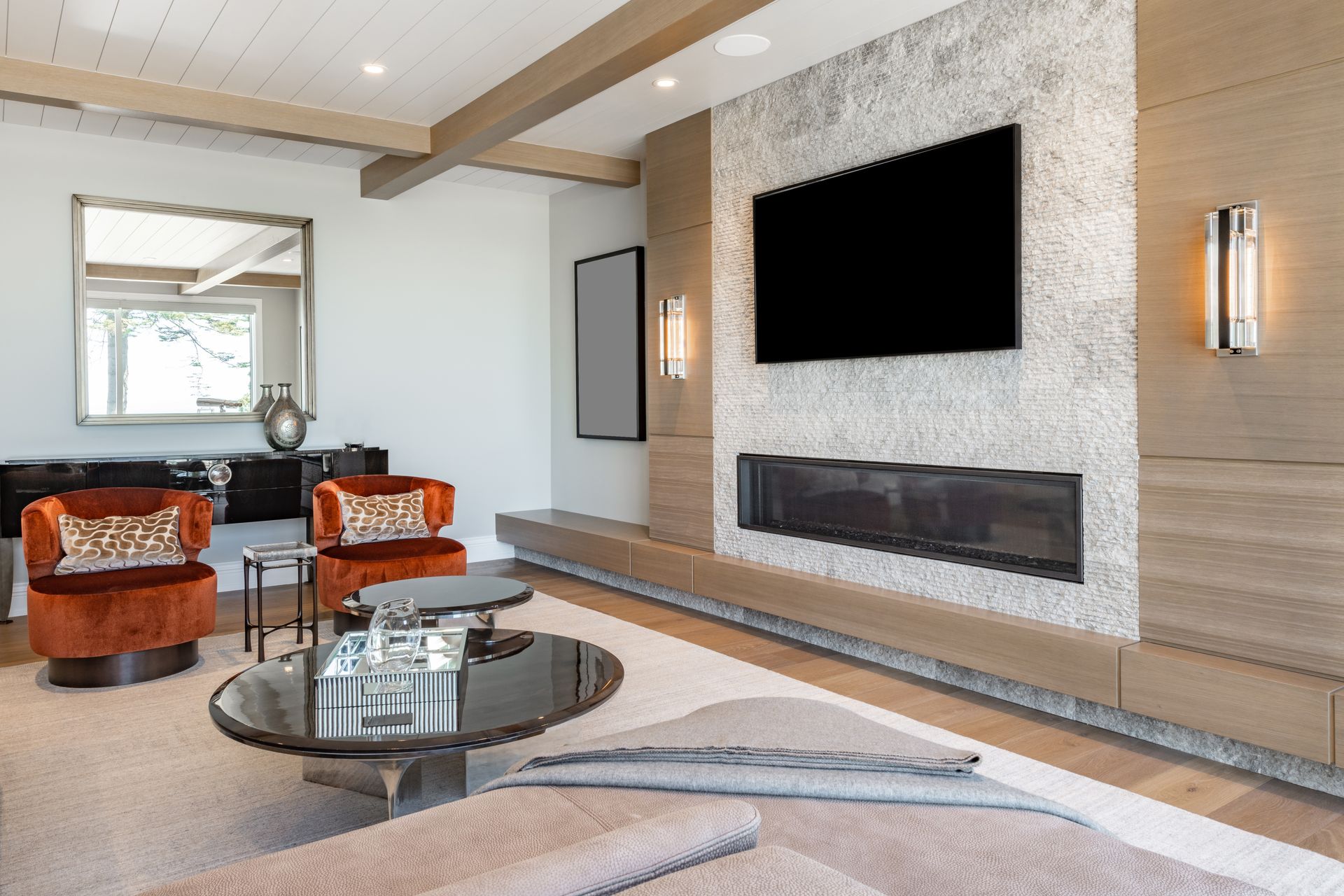 Living room with a large TV above a linear fireplace, two orange armchairs, coffee tables, and a mirror on a white wall.