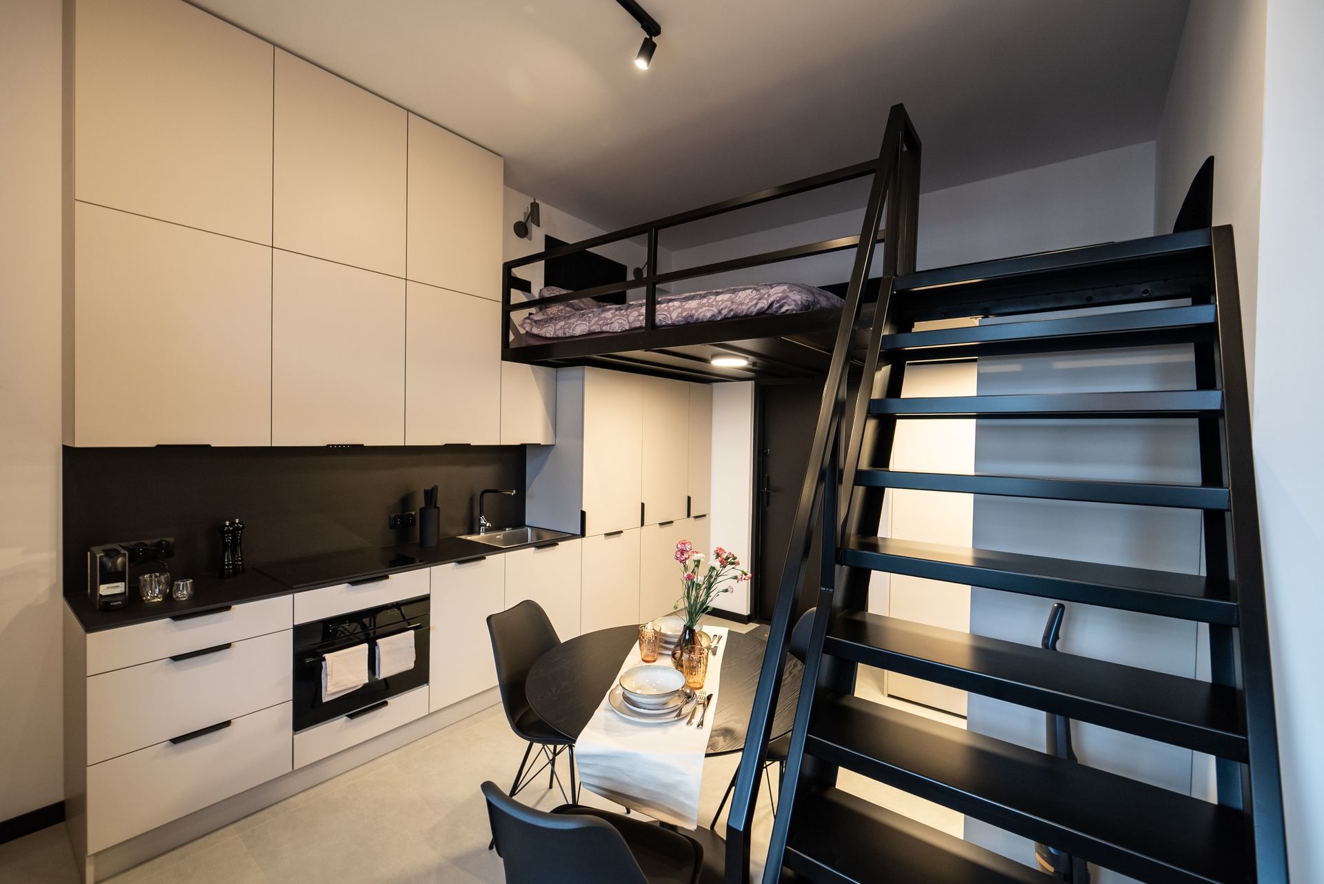 A studio apartment with a loft bed, a black metal staircase, a dining table, and a modern kitchen with light cabinets.