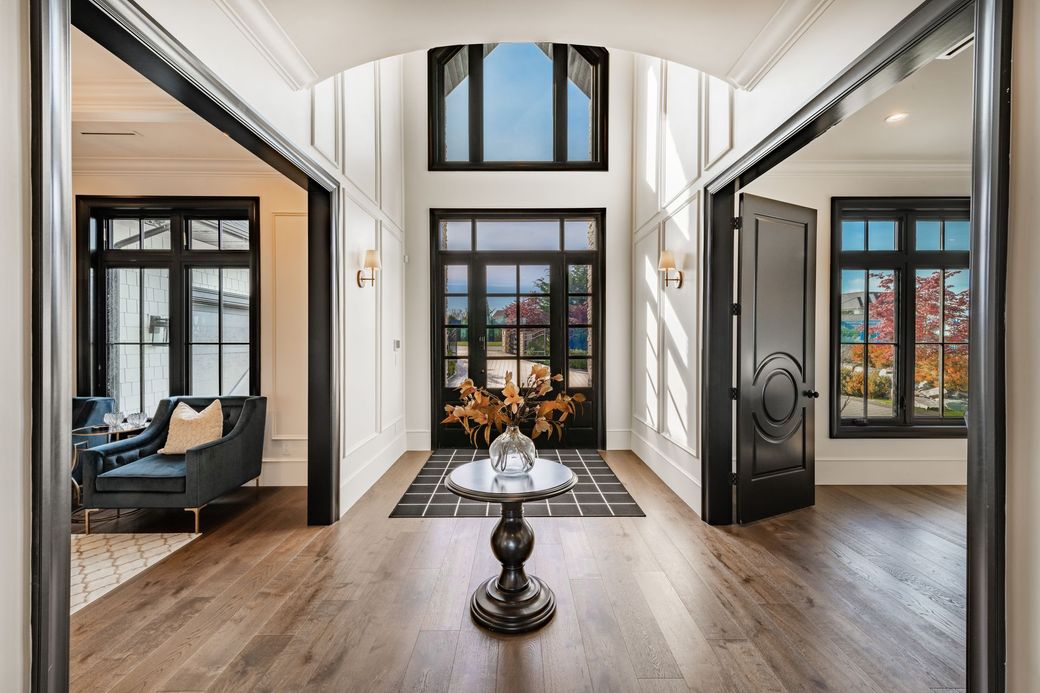 A bright, modern home foyer with a dark arched entryway, hardwood floors, a round pedestal table, and black-framed windows.
