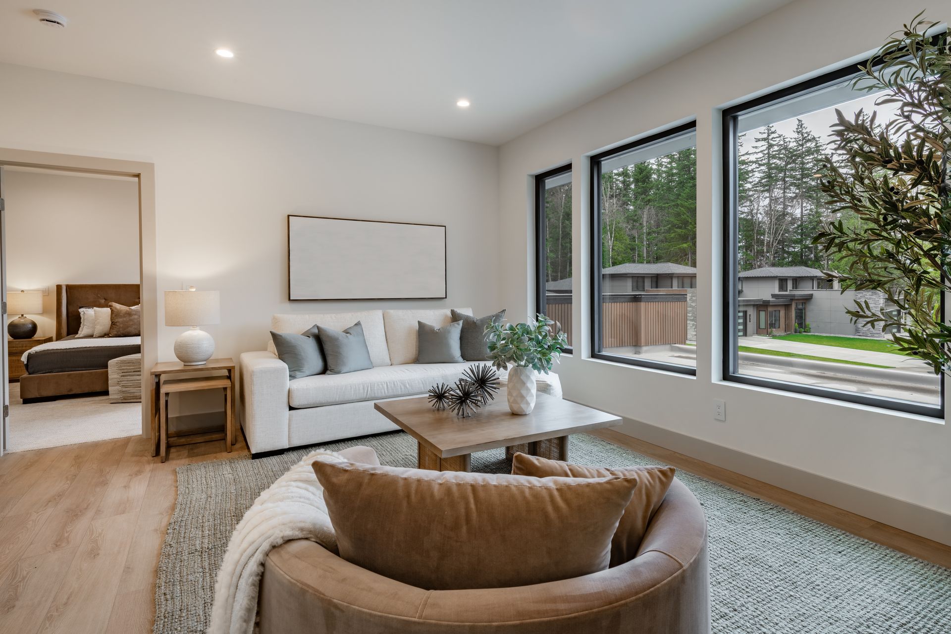 A living room with a white sofa, a tan armchair, a wooden coffee table, and large windows with a view of a neighborhood.